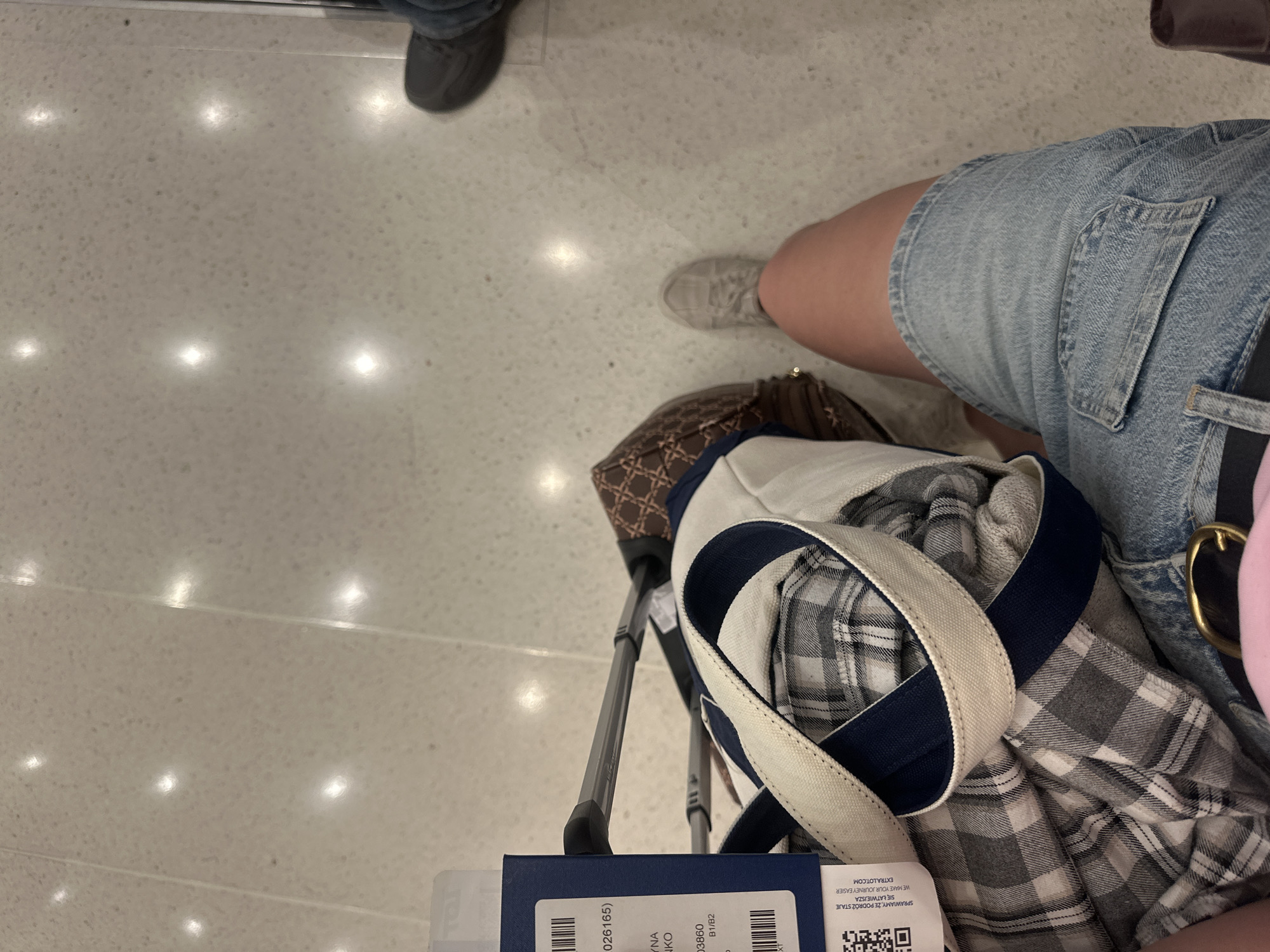 The image shows a person's legs and luggage from a first-person perspective, likely in an airport or train station. The floor is a speckled, light-colored tile with embedded lights. The person is wearing denim shorts and a light-colored sneaker. Next to them is a plaid garment, a canvas tote bag with navy trim, and a suitcase handle. A boarding pass or luggage tag with a barcode and text is visible on the left. No specific landmarks or indications of Chicago are present in the image.