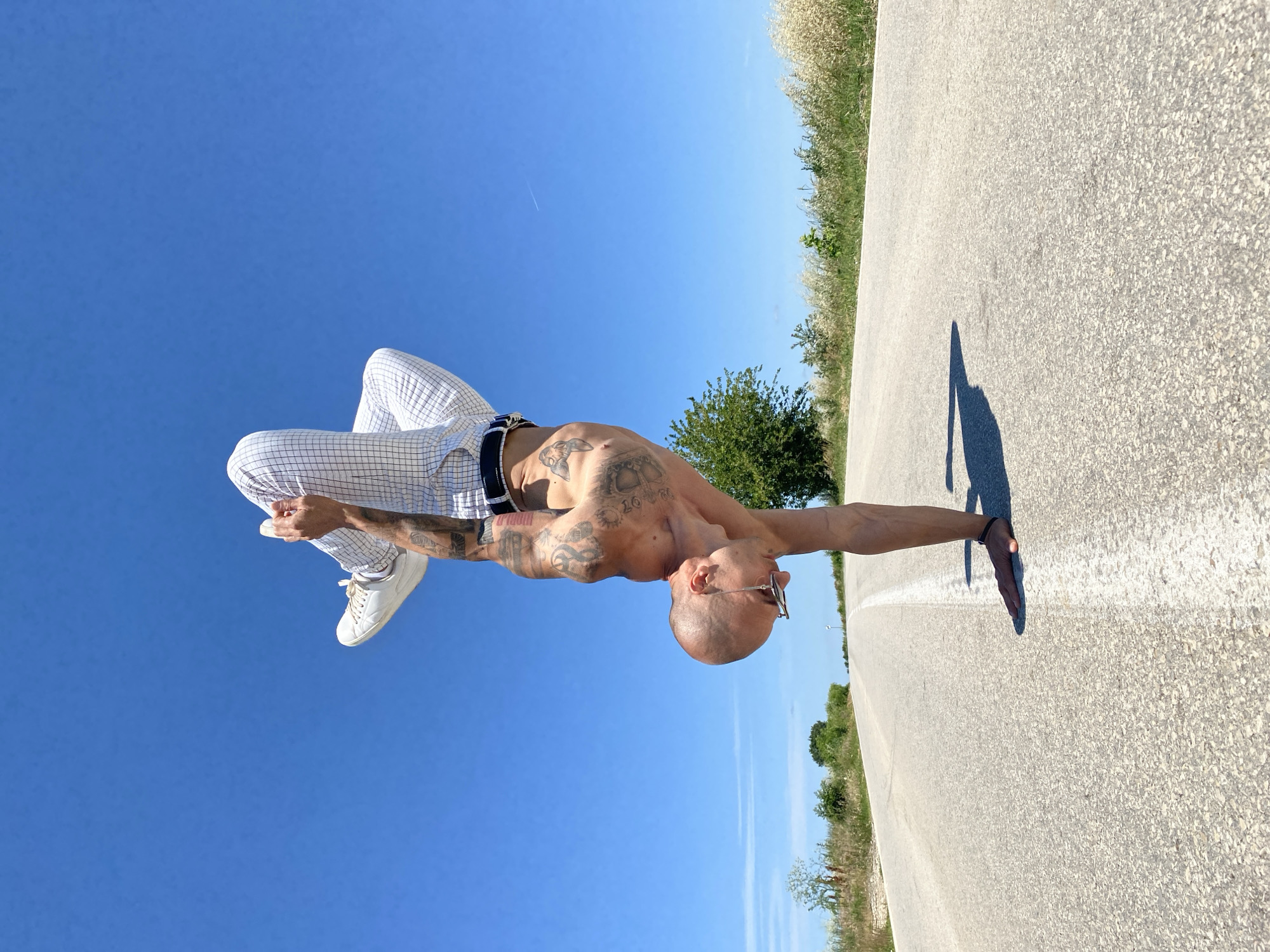 A bald, tattooed man is performing a handstand on a road. He is wearing white checkered pants and white sneakers. His body is inverted, with his feet in the air and his hands on the ground. The road is asphalt and has a white line in the middle. There are grassy fields and trees on either side of the road. The sky is blue and clear. The image was taken in Zgalevo, Bulgaria.