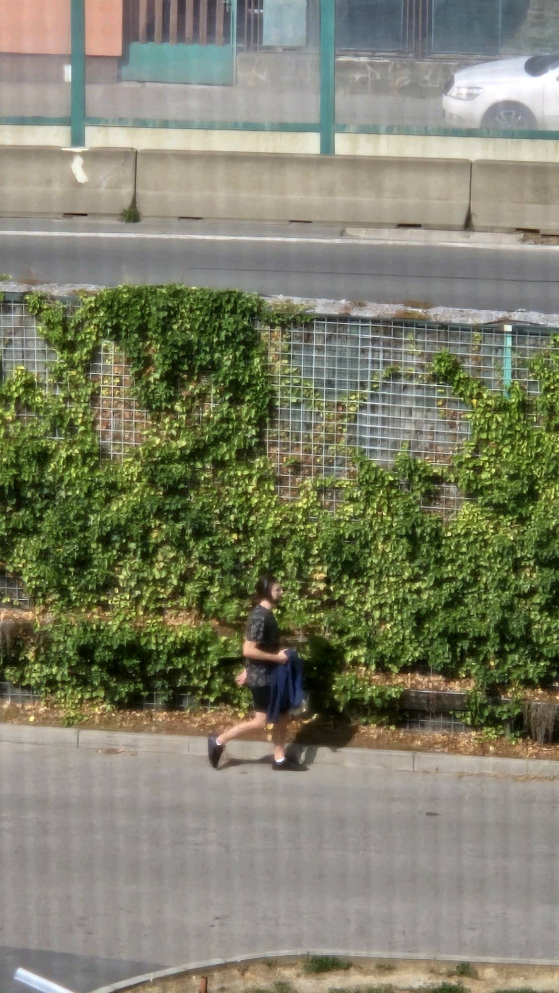 A person with dark hair and a beard, wearing a dark patterned shirt and dark shorts, walks on a paved surface alongside a wall covered in green ivy. The person is holding a dark blue garment in their right arm. A white car is visible in the background, beyond a concrete barrier and a chain-link fence. The scene is outdoors, with evidence of urban infrastructure.