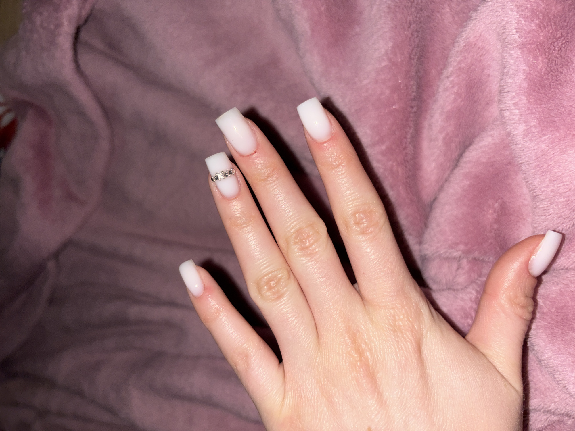 A pale-skinned human hand with white, square-shaped artificial nails rests against a pink, textured fabric. One finger has a line of small, shiny gems or rhinestones adhered to the nail. The location is unknown.