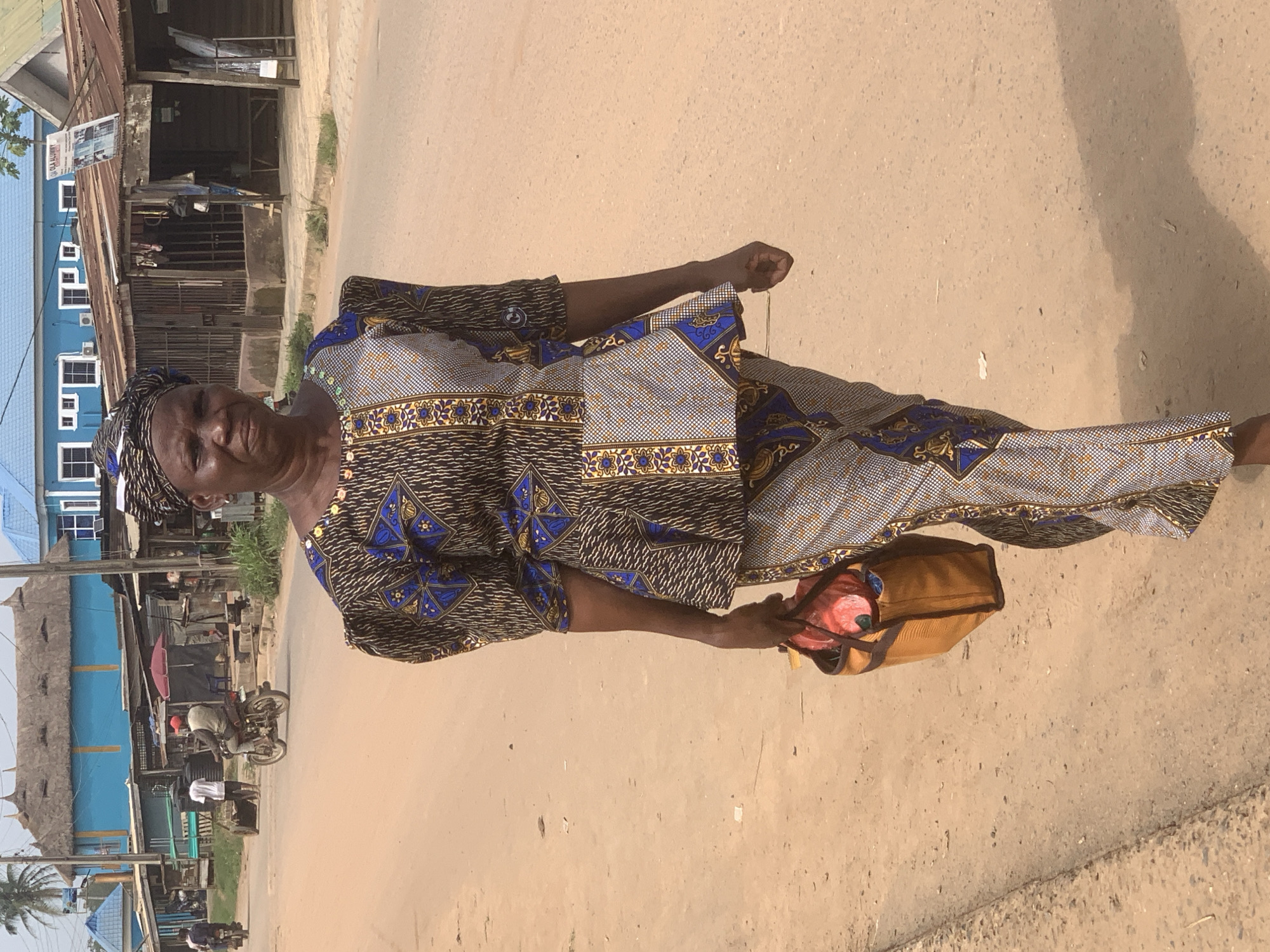 An elderly woman wearing a patterned dress and headscarf is standing on a paved road in Shagamu, Nigeria. She is holding a brown bag in her right hand. Behind her are buildings, some with blue facades and corrugated metal roofs. A motorcycle with a rider is visible in the background on the left. The road surface is uneven and shows signs of wear.