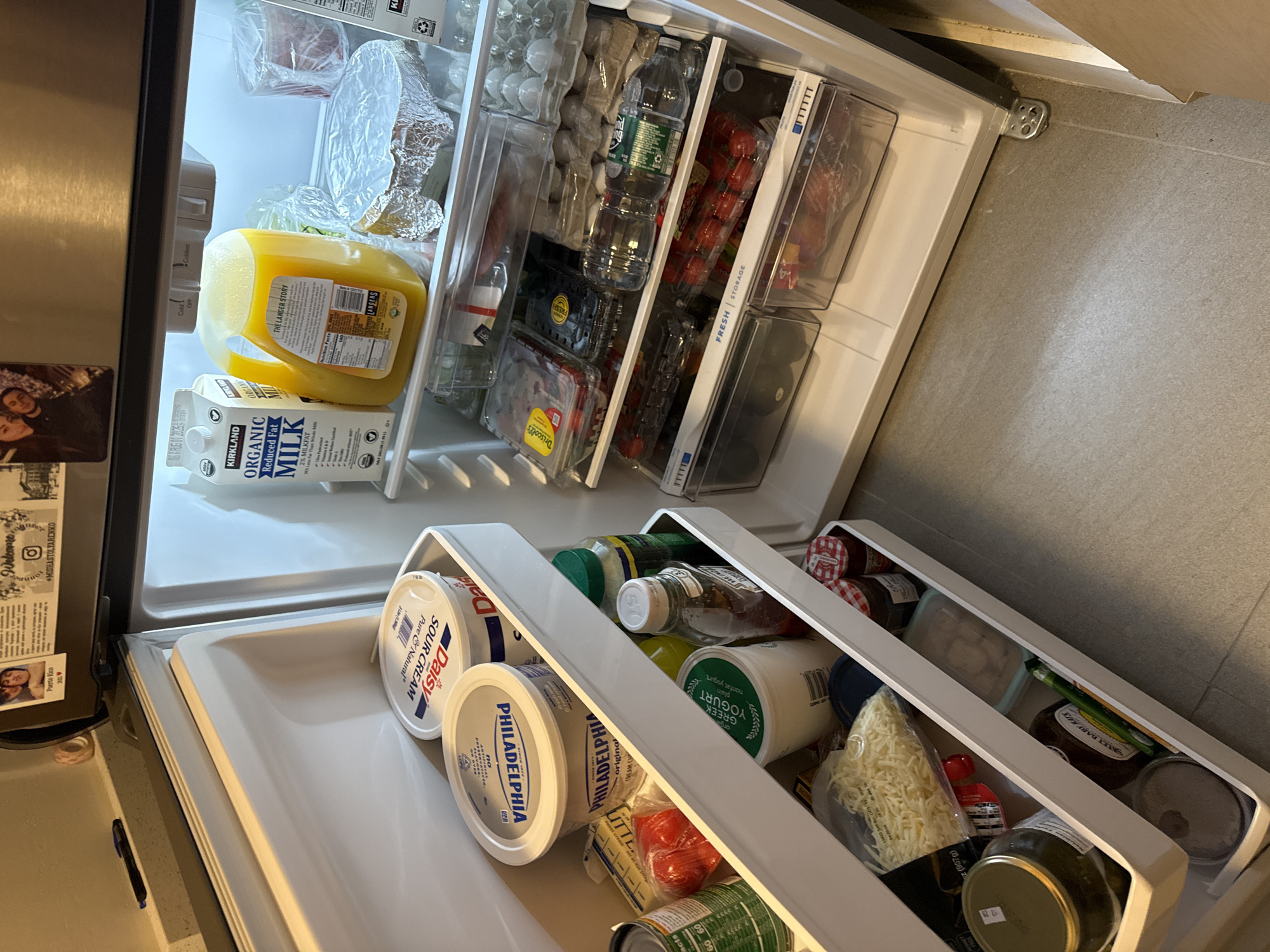 The image shows the inside of an open refrigerator, located on the floor of a kitchen in the City of Yonkers, United States of America. The refrigerator has multiple shelves containing various food items and beverages. On the top shelf, there is a carton of Kirkland Organic Milk and a jug of orange juice. Below that, there are containers of sour cream and cream cheese, along with butter, cherry tomatoes, and canned goods. Further down, there are containers of Greek yogurt, jars of pickles, jam, and barbecue sauce. There are also containers of shredded cheese and various other food items stored in plastic drawers and on shelves. The refrigerator door is open, revealing a well-stocked interior.