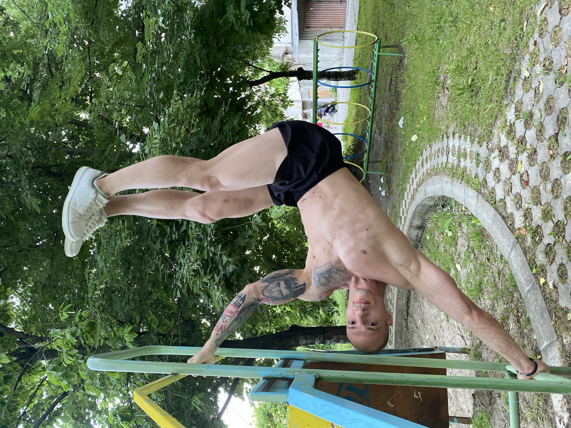 A shirtless man with dark hair, fair skin, and visible tattoos on his left arm is performing an inverted handstand on a multicolored metal playground structure in what appears to be a public park. He is wearing black athletic shorts and white athletic shoes. His body is nearly horizontal, with his legs extended upwards towards a canopy of green trees. In the background, there are green grass areas, a circular paved path with interlocking concrete blocks, and a metal structure with suspended colorful rings. A low building is visible in the distance. The specific location is likely within a park or playground in Pleven, Bulgaria, given the presence of typical urban park infrastructure and playground equipment.