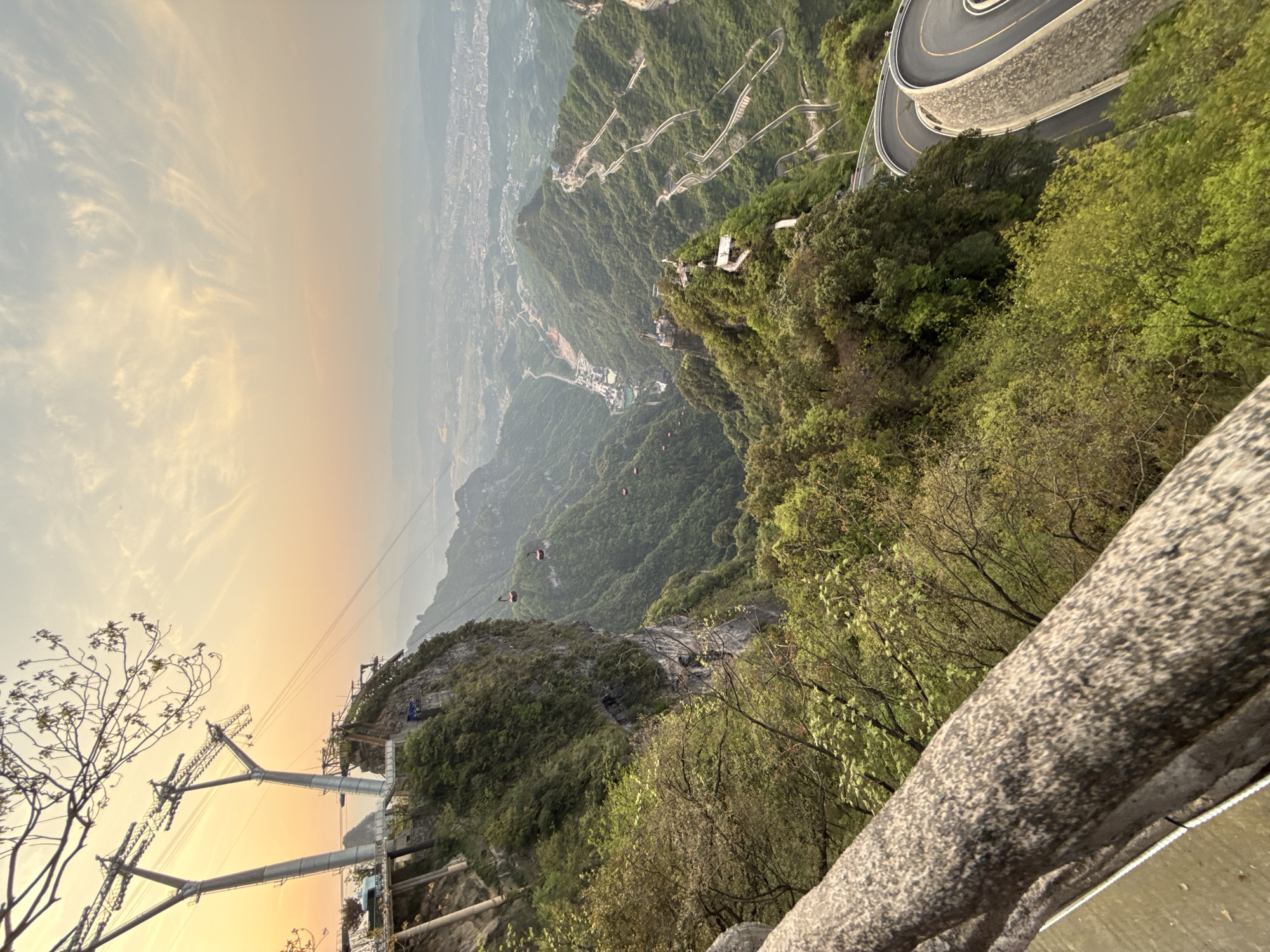 The image shows a panoramic view from a high vantage point in Zhangjiajie, China. The landscape is dominated by lush, green mountains and a valley filled with dense vegetation. In the distance, a city can be seen obscured by a slight haze. A winding road with multiple hairpin turns is visible on the right side of the frame, clinging to the mountainside. A cable car system is present, with several gondolas suspended along the cables, traversing the valley. In the foreground, there's a stone railing, and to its left, the infrastructure of the cable car system, including towers and supporting structures. The sky exhibits a gradient of colors, ranging from a pale yellow-orange near the horizon to a light blue higher up, suggesting either sunrise or sunset.