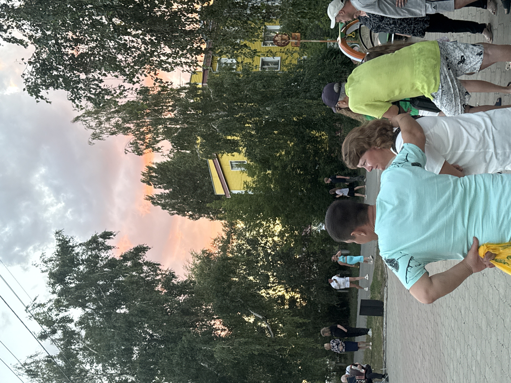 In Yarovoye, Russia, a group of people are gathered outdoors near a bright yellow building with white window frames. The sky above is painted with hues of pink and orange, suggesting either sunrise or sunset, and is partially obscured by the dense foliage of tall trees. In the foreground, a man wearing a turquoise t-shirt with a graphic print stands with his back to the camera, holding a yellow plastic bag in his right hand and resting his left hand on his hip. To his right, a woman with auburn hair looks to her left. Behind them, a man in a neon green t-shirt and patterned shorts stands with his back to the camera. Other individuals are visible further in the background, including a man playing an accordion. A playground structure can be seen to the right of the scene.
