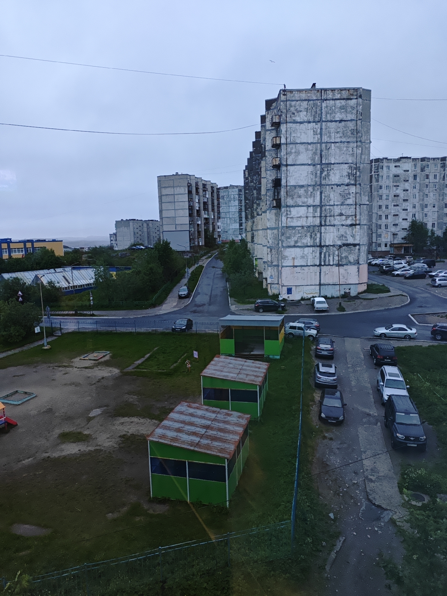 The image shows a cloudy, overcast day in an urban environment with multi-story apartment buildings. In the foreground, there are two green structures with rusty, corrugated metal roofs, possibly shelters or small buildings. Adjacent to these structures is a dirt and grass area with a children's sandbox. A dark fence runs along the bottom edge of the frame. To the right, a narrow, paved road curves upwards, lined with parked cars, including a silver sedan, a black SUV, and several other vehicles. Further up the road, a large, weathered, multi-story apartment building with a gray facade dominates the view. Other apartment buildings, also appearing to be of similar construction and age, are visible in the background. A greenhouse-like structure with a glass roof is situated to the left of the road, surrounded by trees. The overall impression is one of a somewhat dilapidated, residential area under a gray sky.