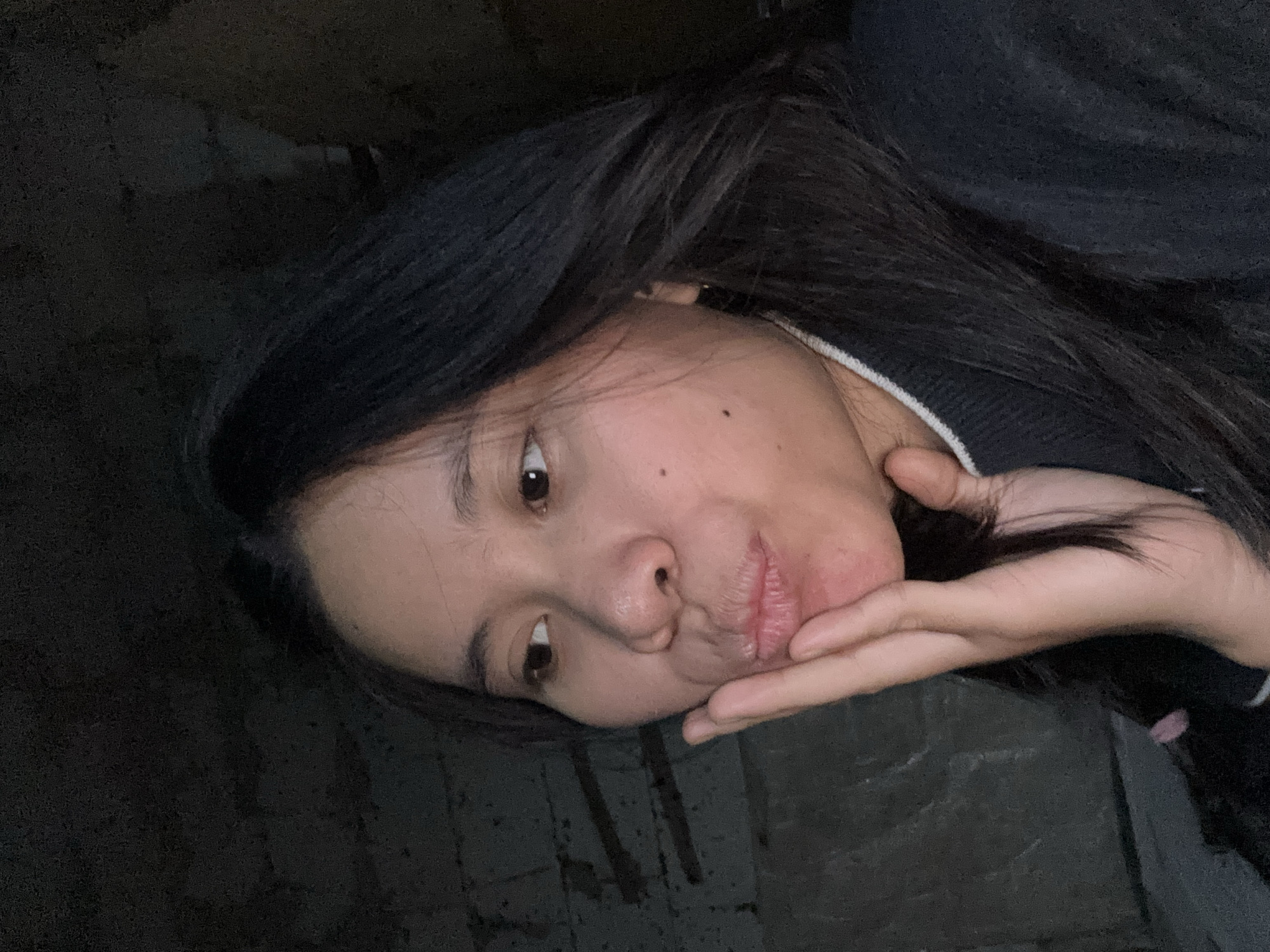 In a dimly lit, indoor environment, a young woman of East Asian descent is captured in a close-up, selfie-style photograph. She has long, dark hair and is wearing a dark-colored sweater with a white collar. Her left hand is raised, with her fingers gently touching her cheek and lower lip, and her lips are pursed in a pout. Her eyes are directed to her left, and she has a small mole on her right cheek. The background appears to be a rough, textured wall, possibly brick, with some wooden elements or debris visible. The overall lighting is low, casting shadows and creating a somewhat somber atmosphere. The location is specified as East Jakarta, Indonesia.