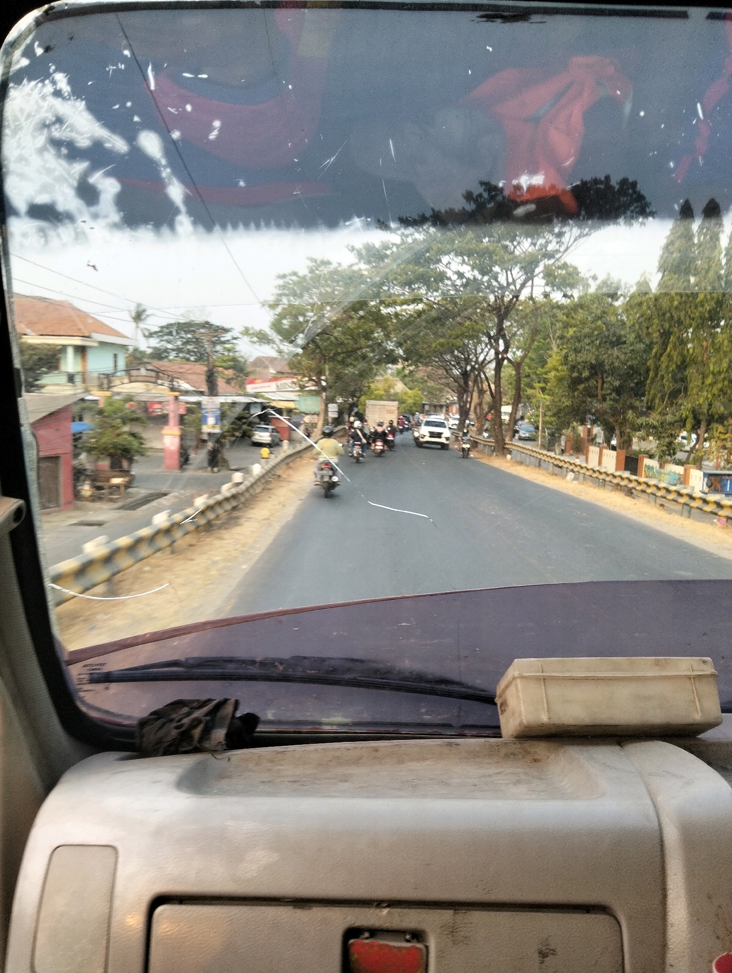 The image shows a view from inside a vehicle, looking out through the windshield. The windshield is cracked and smudged, with a reflection of what appears to be red fabric in the upper portion. In the foreground, on the dashboard, is a crumpled black cloth and a light-colored rectangular box.
Outside the vehicle, a two-lane road is visible, with traffic moving in both directions. Several motorcycles and a white car are on the road. A yellow and black striped guardrail lines the right side of the road, separating it from a dirt embankment. Beyond the embankment, trees and buildings line the street. The buildings appear to be single or two-story structures, with some displaying signs that suggest commercial activity. The overall scene suggests a moderately busy street in an urban or semi-urban area.
Based on visual cues such as the architecture of the buildings and the general ambiance, the scene is likely located in Semarang, Indonesia. The presence of numerous motorcycles, the style of the vehicles, and the tropical vegetation are all consistent with this location. The time of day appears to be daytime, possibly late afternoon, given the lighting and the shadows.