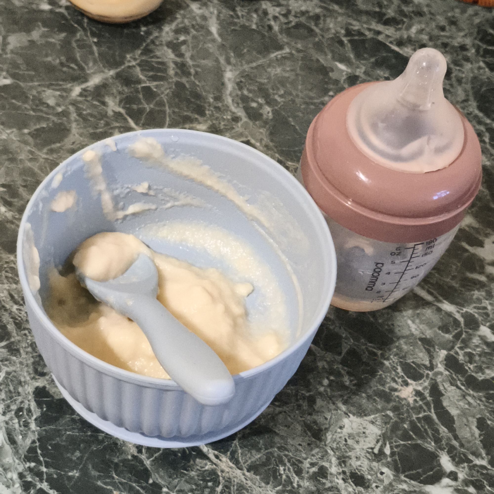 A close-up shot shows a baby bottle and bowl of baby food on a marble countertop. The bowl, light blue in color, contains a pale yellow pureed food with a spoon resting inside. The baby bottle is partially filled with a white liquid. The nipple is clear, and the bottle is marked with measurements in milliliters and ounces. The bottle's cap is pink. The background consists of a marble surface with dark green and white veining.