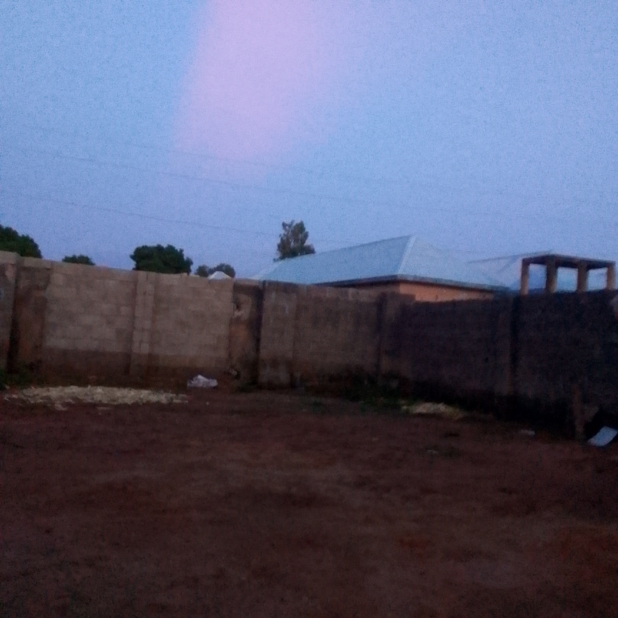 In the city of Unknown, Unknown, at dusk, a dirt ground is visible in the foreground. Beyond the dirt, a weathered concrete block wall runs horizontally across the image, extending from left to right. On the left side of the wall, there are piles of light-colored straw or vegetation. Behind the wall, in the middle ground, is a structure with a light blue metal roof, possibly a house or building, with an orange-colored wall visible. To the right of this structure, there is another partially constructed concrete structure with visible columns. Trees with dark green foliage are situated behind the wall and to the left and right of the buildings. The sky above is a gradient of pale blue and pink hues, indicating either sunrise or sunset. No people are present in the scene.