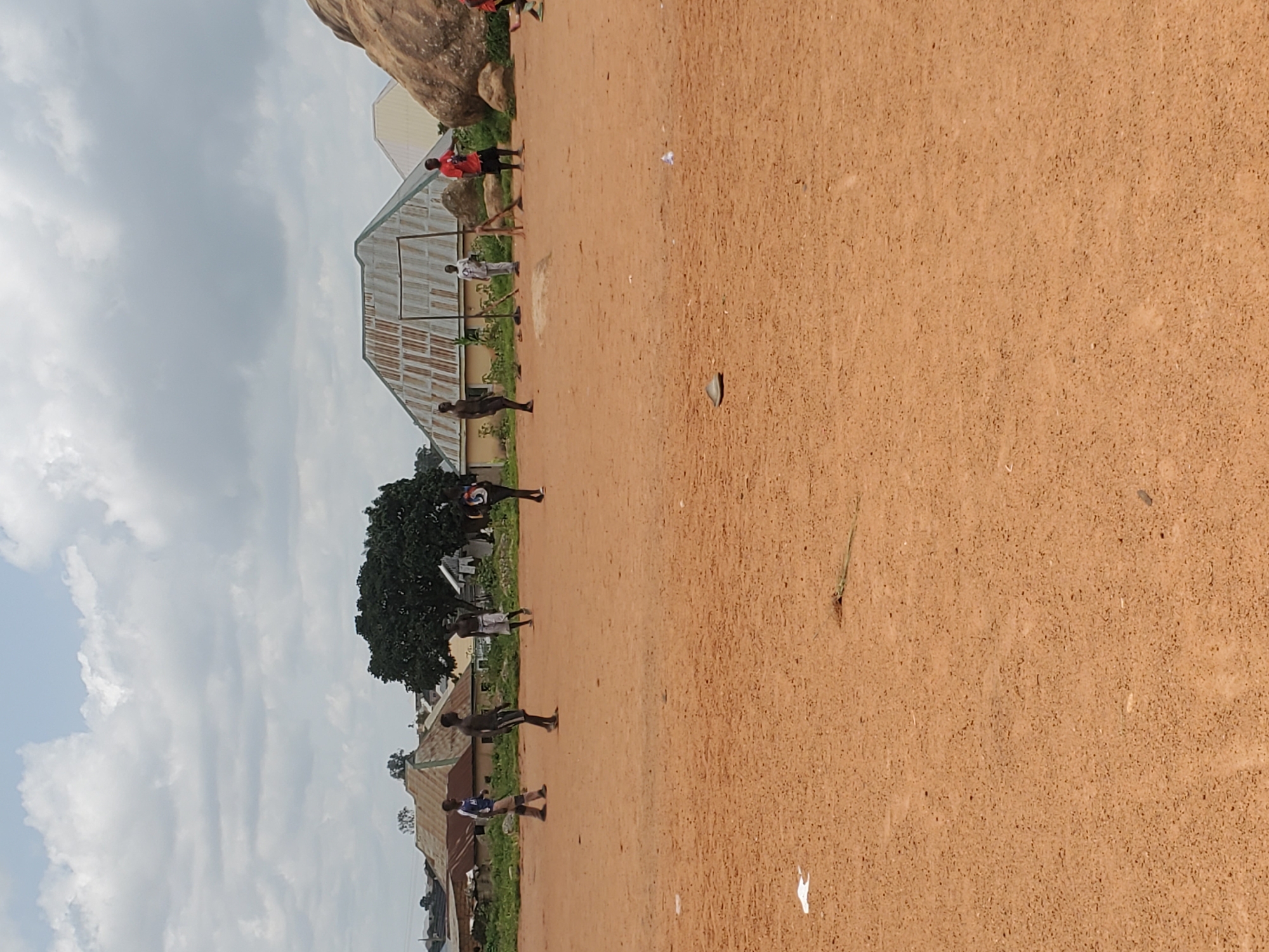 In Jos, Nigeria, on a sandy, uneven field, several boys are engaged in a game of soccer. The foreground is dominated by the reddish-brown earth of the playing surface, with a few scattered pieces of debris, including a white plastic bag and a dark, flat object. In the middle ground, the boys are spread out, some actively participating in the game, kicking or running, while others observe. A makeshift goal, constructed from metal poles and wooden supports, is visible. The background features a collection of houses with corrugated metal roofs, indicative of a residential area. Lush green trees and a large rock formation on the right side of the image provide a natural element to the urban landscape. The sky above is partly cloudy with a mix of white and grey formations.