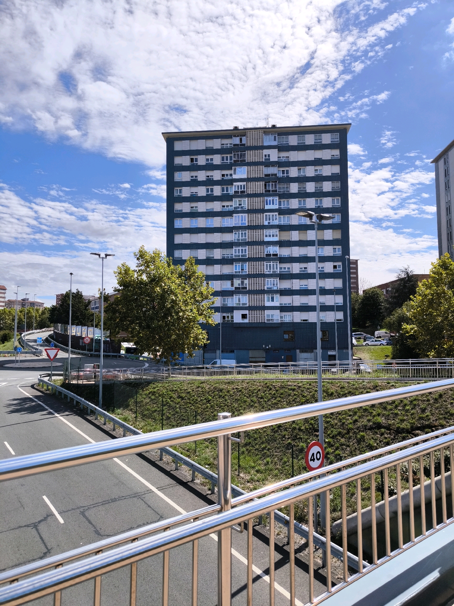 The image depicts a daytime scene in an urban environment, likely Barakaldo, Spain, characterized by a large, dark teal apartment building with numerous windows and white balconies occupying the central and right portions of the frame. The building has a flat roof and appears to be several stories high. In front of the building, a mature, leafy green tree stands prominently. To the left of the apartment building, a curved roadway with white lane markings is visible, bordered by a metal railing. A red and white speed limit sign displaying "40" is attached to a pole near the roadway. The sky is blue with scattered white clouds, and the ground is covered in green grass and some patches of darker soil. Further in the background, other buildings and trees are discernible, suggesting a residential or mixed-use urban area. There are no people or discernible actions taking place in the image.