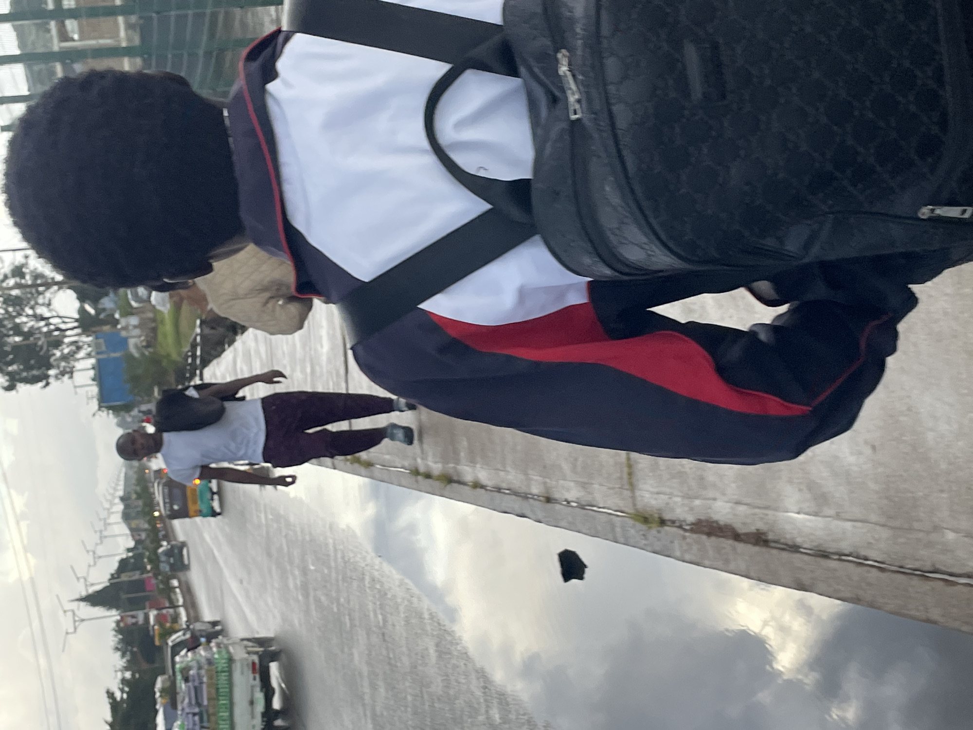An overhead perspective shot captures two individuals walking on a sidewalk next to a wet road. The person in the foreground, facing away from the camera, wears a white, red, and blue jacket over a white shirt, and a black patterned backpack. To their left, a man in a white t-shirt and dark pants walks away from the camera, carrying a dark jacket over his left shoulder. The road appears to have a large puddle reflecting the cloudy sky. In the background, a white truck loaded with green boxes is visible, along with a yellow and green rickshaw. Further down the road, cars and other vehicles can be seen. The environment suggests an urban setting, possibly in Barakin Lerawi, Nigeria, with streetlights and overhead wires lining the road. The overall lighting indicates it is daytime, possibly late afternoon, with overcast skies.