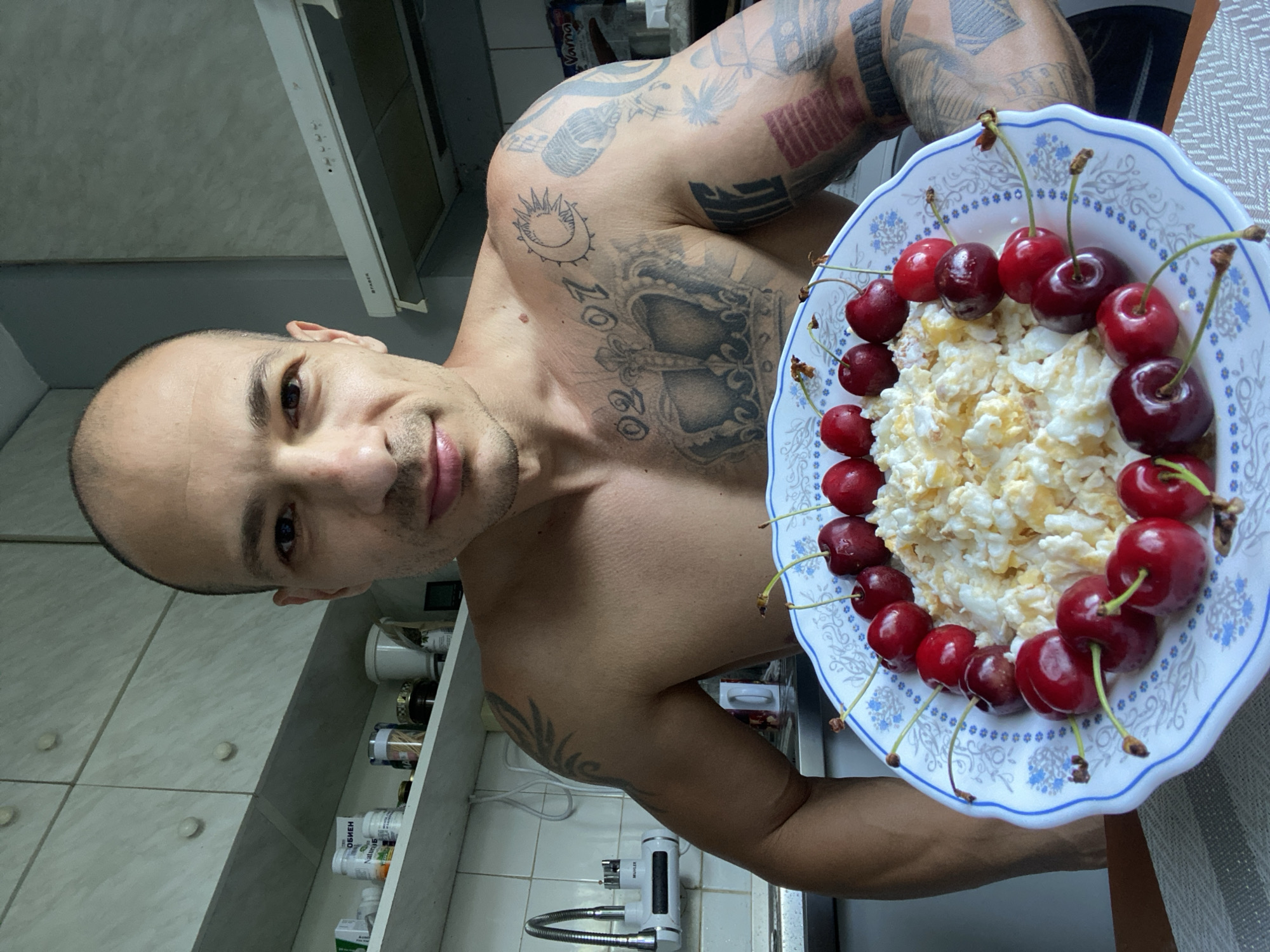 A shirtless man with tattoos on his chest and arms is holding a white plate with a blue floral pattern. The plate contains a mound of scrambled eggs topped with a ring of cherries. The man is in a kitchen with light-colored cabinets and a tiled backsplash. A faucet is visible on the left side of the image, and a range hood is above the stove. The overall setting is casual and domestic. The image was taken in Pleven, Bulgaria.