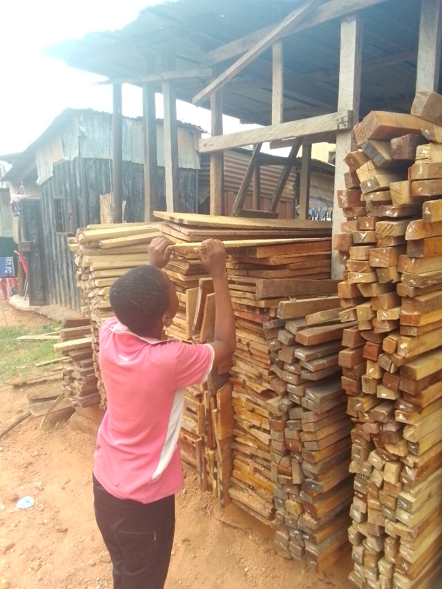 In an outdoor scene, a person is shown from behind, actively engaging with tall stacks of lumber under what appears to be an open-sided shelter. The individual wears a pink short-sleeved shirt with white accents and dark pants, their hands placed on top of a pile of wooden planks, suggesting they are in the process of stacking, organizing, or moving the wood.
The setting is a lumber yard or sawmill area, likely in Onitsha, Nigeria, characterized by reddish-brown dirt ground. Multiple large stacks of wooden planks, varying in size and hue, dominate the midground and right side of the image. The wood appears to be freshly cut or prepared, with some planks showing end grain markings.
In the background, several structures made from corrugated metal sheets and wooden frames are visible, indicating an informal or industrial area. One building features what looks like laundry hanging on a clothesline. The person and the lumber are beneath a basic wooden framework supporting a roof, providing shelter.
The lighting is bright and even, suggesting daytime, possibly mid-morning or afternoon, with no harsh shadows. The sky is largely overexposed in the upper left, contributing to the bright atmosphere. On the far left, a red object with partially visible text, possibly "EA FC," can be faintly discerned. A small blue plastic item lies on the dirt ground near the person's feet. The overall scene depicts labor related to the timber industry in a functional, open-air environment.