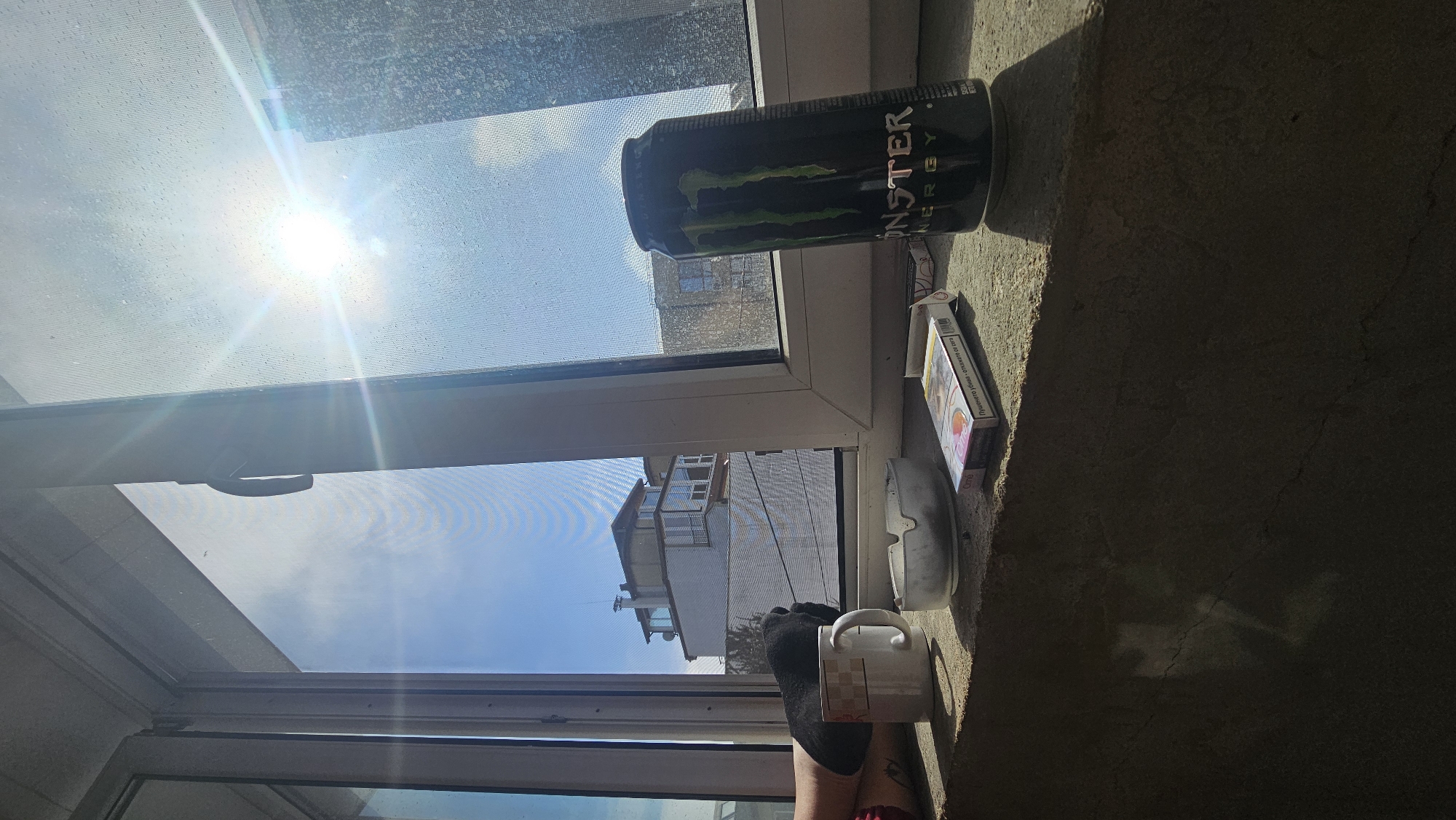 The image depicts a view from a window in Varna, Bulgaria, judging by the architectural style of the buildings visible. A partially opened window frame is prominent, allowing a view of a bright sky with the sun shining through. On the window sill, there is a can of Monster Energy drink, a pack of cigarettes, a white ashtray, a white mug, and a leg covered with a black sock. The windowsill is made of rough gray stone. The leg appears to be resting near the mug, suggesting a casual or relaxed setting. The scene conveys a sense of domesticity combined with common consumer products.