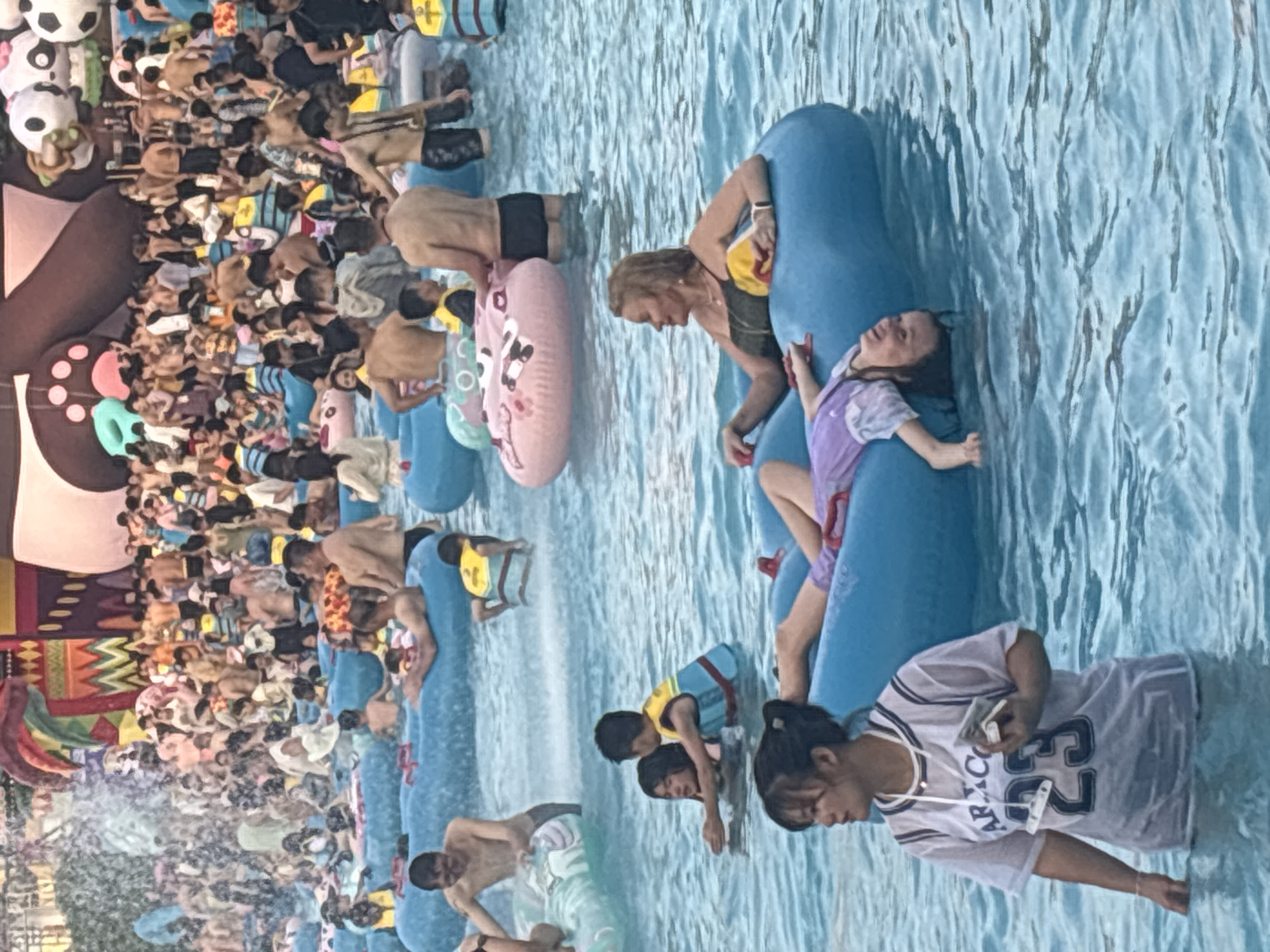 This image depicts a crowded scene at a water park, possibly in Panyu District, China, given the context. The water is a clear, artificial blue, with ripples indicating movement. Numerous people are engaged in water-based activities, with many floating on inflatable tubes. The overall atmosphere appears to be one of leisure and recreation.
In the foreground, a young woman wearing a translucent white t-shirt over a dark swimsuit stands partially submerged in the water. She holds a small, rectangular object, possibly a phone or wallet, in her right hand and has a necklace around her neck. To her left, a child with dark hair is lying on a blue inflatable float, wearing a purple outfit. Her head is tilted back, and her eyes are closed, suggesting relaxation or sleep.
Further into the scene, a woman with blonde, shoulder-length hair is seated on a blue inflatable tube. She is wearing a dark, strapless swimsuit and a necklace. Her right hand rests on the tube, and she looks to her left. Behind her, a boy wearing a yellow and blue life vest is visible in the water.
The middle ground of the image is filled with a large number of people, many of whom are also on inflatable tubes. Some are standing or wading in the water, while others are seated or lying down. The water is a vibrant blue, and the people are diverse in age and attire, with many wearing swimwear and life vests. In the background, a large, colorful inflatable structure, possibly a slide or a themed decoration, is visible, featuring cartoonish elements like large eyes and paws. The crowd suggests a popular attraction, likely a water slide or a wave pool area. The lighting suggests it is daytime, with bright, diffused light illuminating the scene.