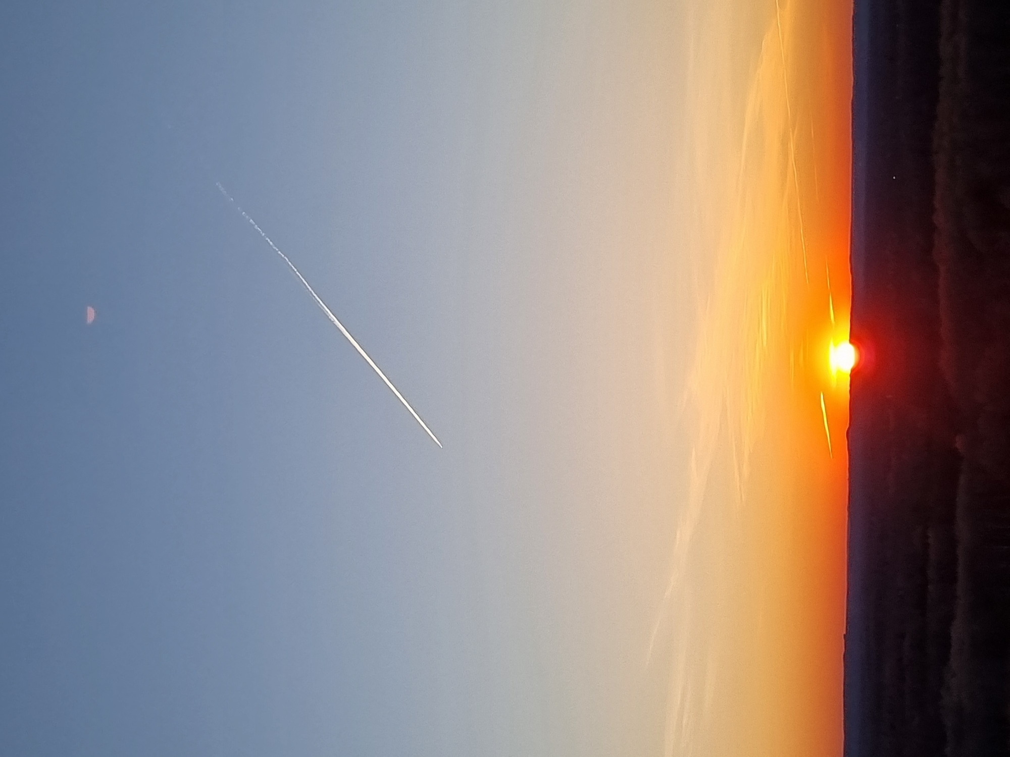 The image depicts a sunset sky. The right side of the image shows a fiery orange and yellow gradient transitioning to a dark, possibly treeline-obscured, horizon. The sun is visible as a bright red-orange orb. A contrail stretches across the blue and lighter orange portion of the sky, moving from upper left to lower center. A faint, small, red circular shape, possibly the moon, is in the upper left of the image. The location, Kemul, Russia, cannot be determined from this image.