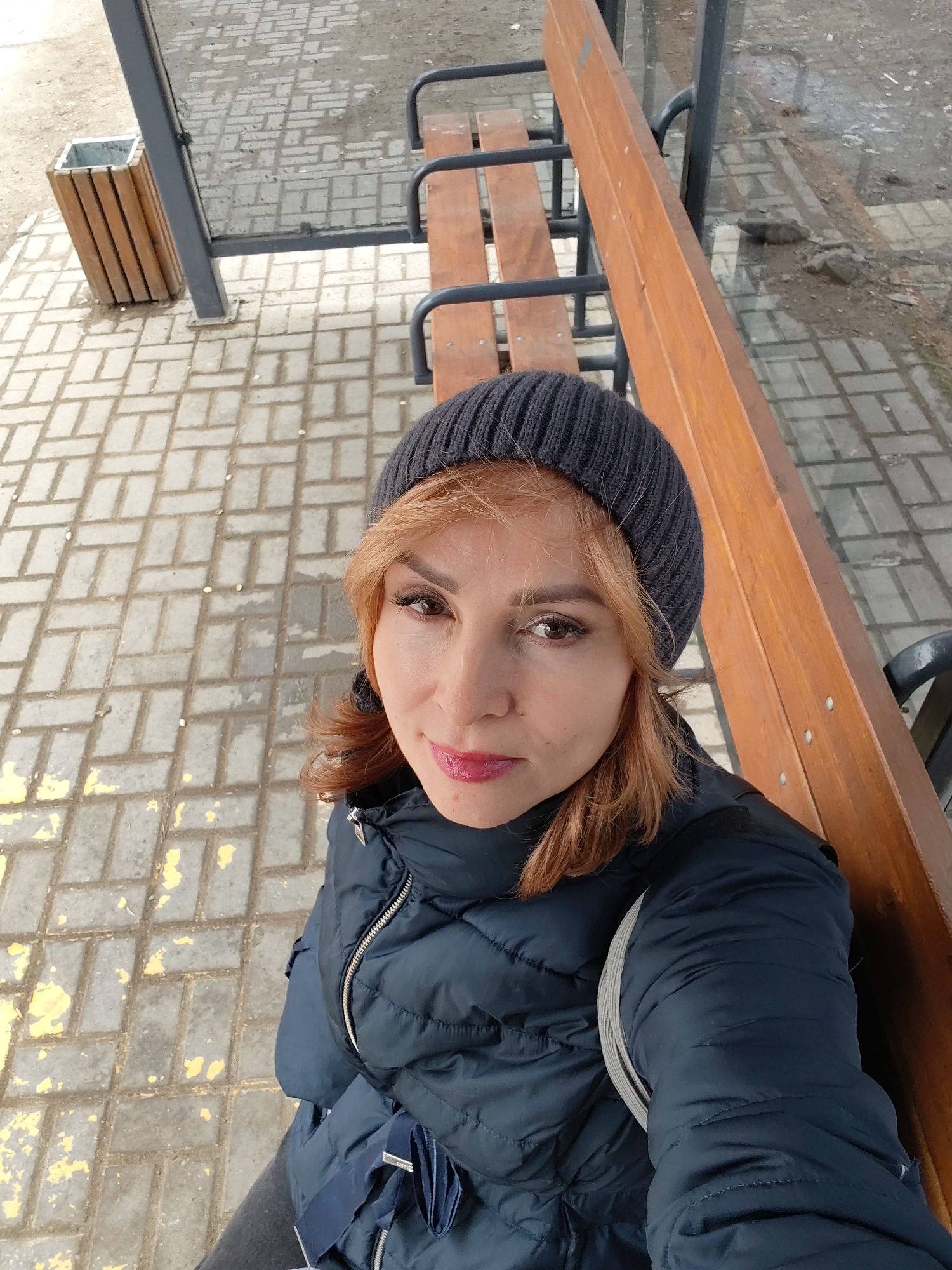 A woman sits on a wooden bench at a bus stop in Chaikovsky, Russia. The bus stop has a gray metal frame and a wooden trash can. The ground is paved with gray bricks with yellow markings. The woman is wearing a dark blue puffer jacket and a gray beanie. She has red hair.