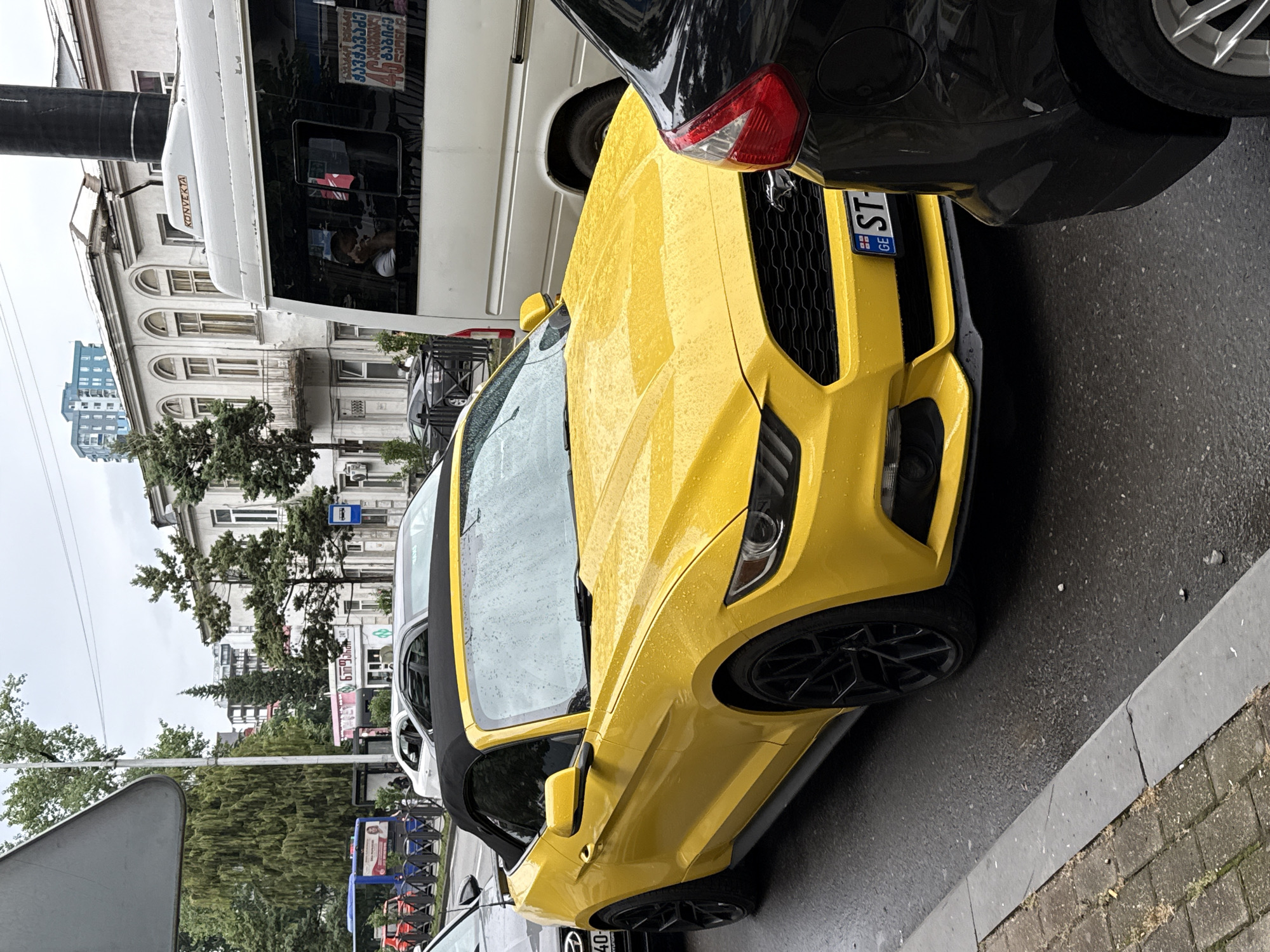 A bright yellow Ford Mustang convertible is parked on a wet city street, its black top up. The car is positioned in the foreground, angled slightly towards the left. Raindrops are visible on the car's surface and windshield. To the right of the Mustang, the rear of a black car is visible. Behind the yellow car, a white van and an older, light-colored building with arched windows and a balcony can be seen. A blue bus and other vehicles are visible further down the street, alongside trees and other buildings. The street appears to be paved with asphalt, and a concrete curb separates the street from a sidewalk made of interlocking bricks. The sky is overcast, suggesting a cloudy or rainy day.