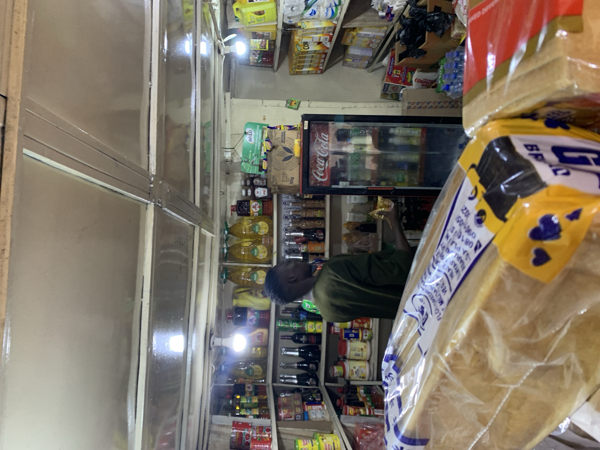 A person in a dark green shirt is standing in a small grocery store. The store is filled with shelves of various products, including bottled drinks, canned goods, and bottled oils. A Coca-Cola refrigerator is in the middle of the back wall. In the foreground, a loaf of bread in a plastic bag obscures the view. Based on the OCR and other details, it is likely this store is located in or near New Stadium, Nigeria.
