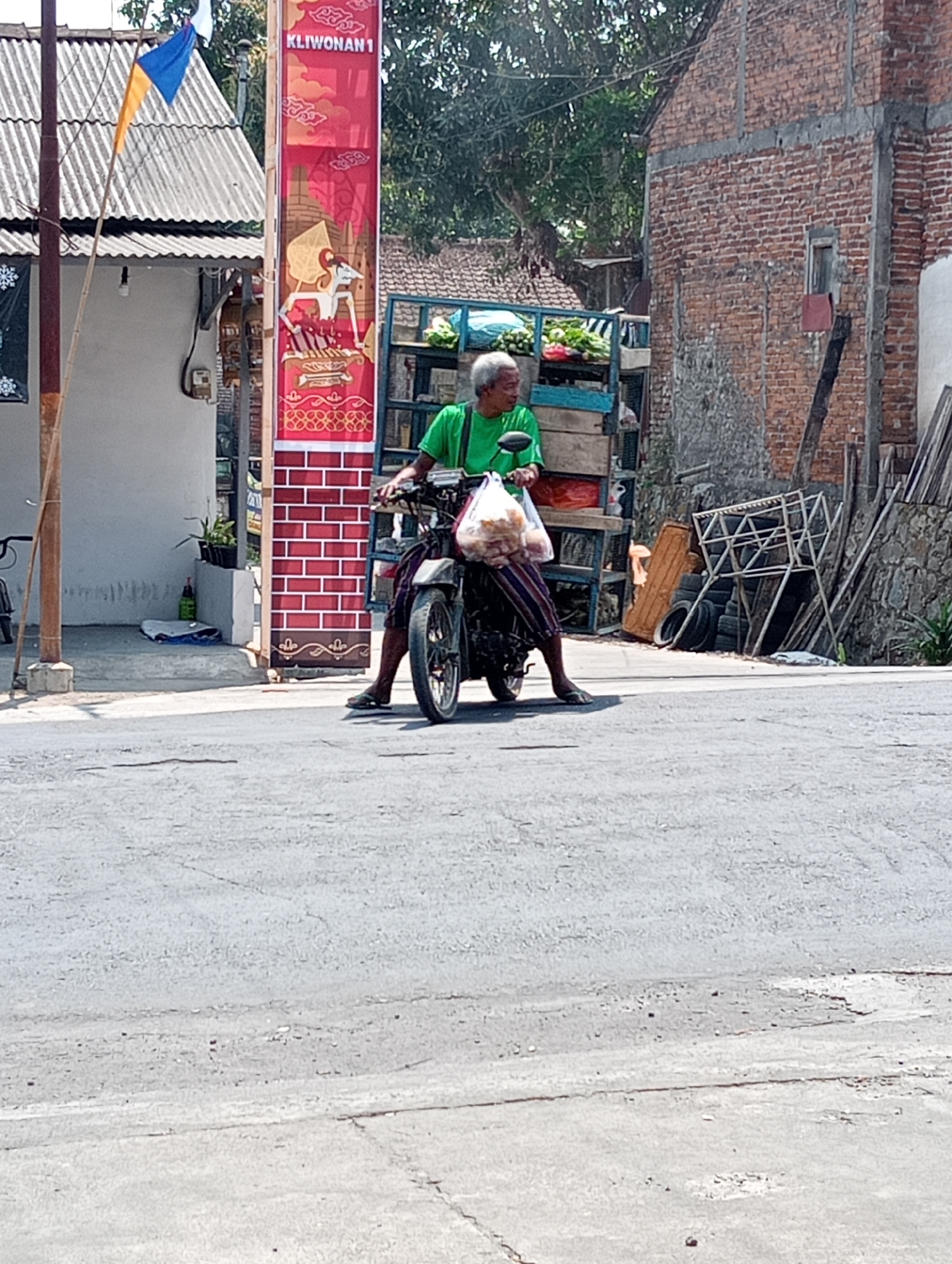In the city of Grabag, Indonesia, a person is riding a motorcycle on an asphalt road. The person, who appears to be an elderly man with grey hair, is wearing a green t-shirt and patterned pants. He is carrying a plastic bag filled with bread in front of him. The motorcycle is dark colored and has a license plate visible.
The background shows a mix of buildings and vegetation. To the left, there is a building with a corrugated metal roof and a banner with red and gold patterns that reads "KLIWONAN 1". A flag with blue, yellow, and white colors is also visible. To the right, there is a brick building with exposed concrete elements. A stall with blue and wooden shelves, filled with produce, is positioned behind the motorcycle. Further to the right, there are stacks of tires and wooden materials. Trees are present in the background, indicating a semi-urban or rural setting. The scene is illuminated by daylight.