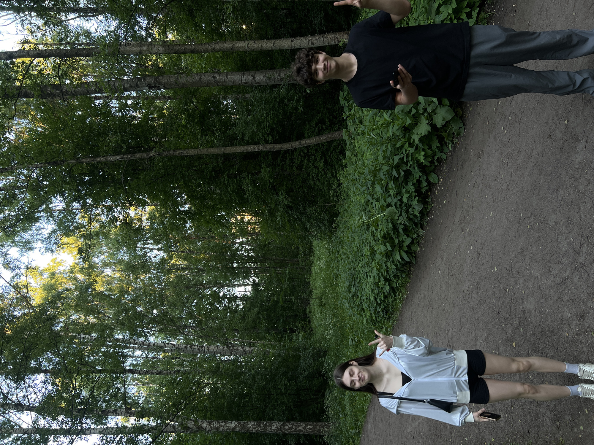 Two young adults, one female and one male, stand on a dirt path within a densely wooded park in Saint Petersburg, Russia. The female, positioned on the left, has long brown hair and wears a white zippered hooded jacket over a black top, black shorts, white socks, and light-colored sneakers. She holds a dark smartphone in her left hand and makes a "rock on" hand gesture with her right hand, her eyes closed and a slight smile on her face. The male, positioned on the right, has short, curly brown hair and wears a black t-shirt and grey track pants. He holds a dark smartphone in his left hand and makes a "peace" sign with his right hand, smiling directly at the camera.
Both individuals are situated on a wide, unpaved dirt path that narrows as it extends into the background. The environment is characterized by a dense forest comprising numerous tall, slender trees, many exhibiting the distinctive white and black bark of birch trees, alongside others with darker, rougher bark. A thick undergrowth of various green plants borders both sides of the path. Sunlight penetrates the tree canopy from above and slightly behind, illuminating the upper foliage with a golden hue. The overall lighting suggests either late afternoon or early morning. The setting appears to be a natural, forested park, typical of the larger green spaces found in Saint Petersburg.