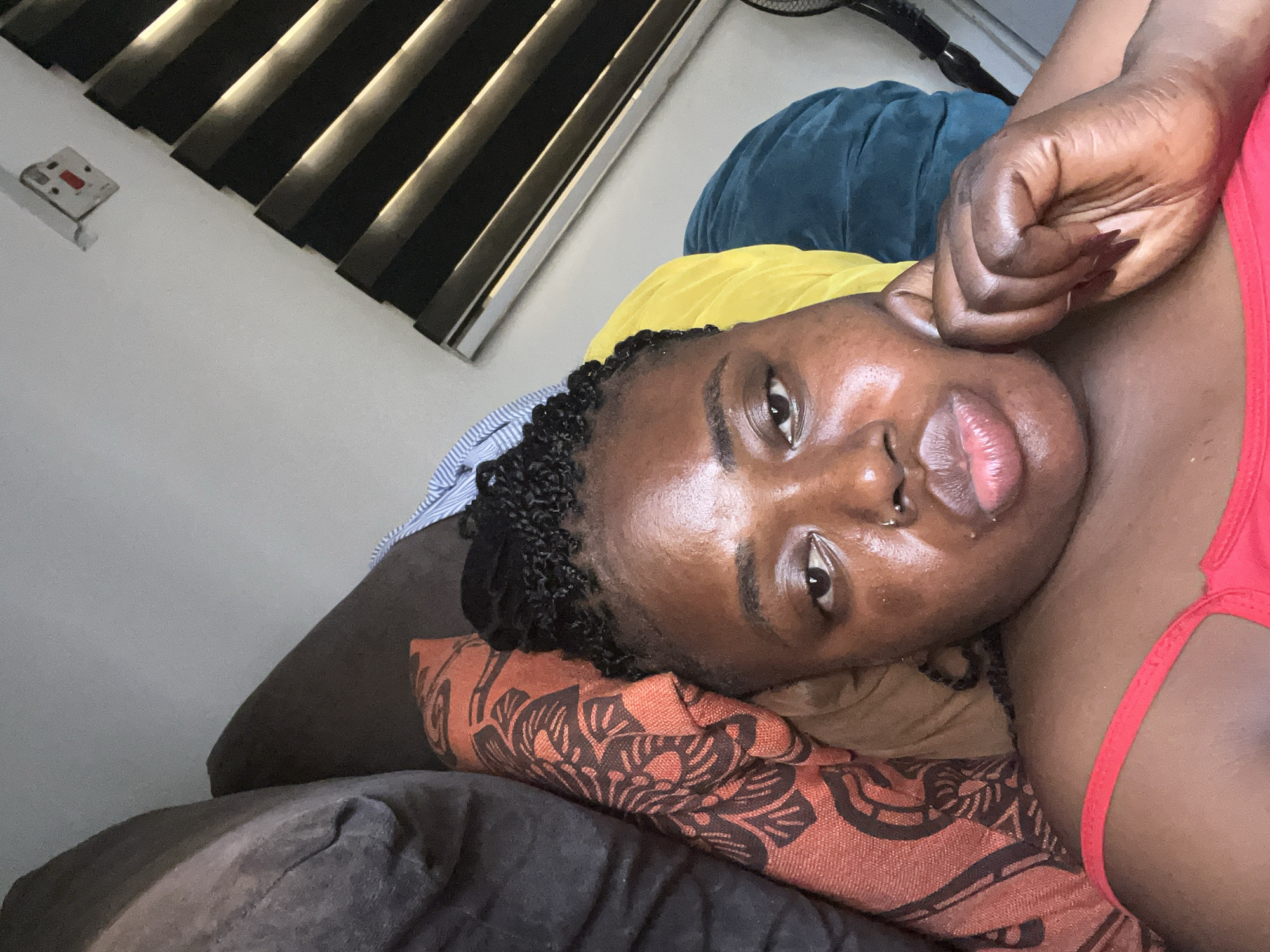 In a bedroom in Abuja, Nigeria, a young Black woman is lying on her side on a sofa, resting her head on pillows and blankets. She is wearing a red tank top. Her hair is braided and tied up. She has a nose ring. Her left hand is resting on her cheek. The room is lit by natural light coming from blinds on a window, and a power outlet with a switch is visible on the wall.