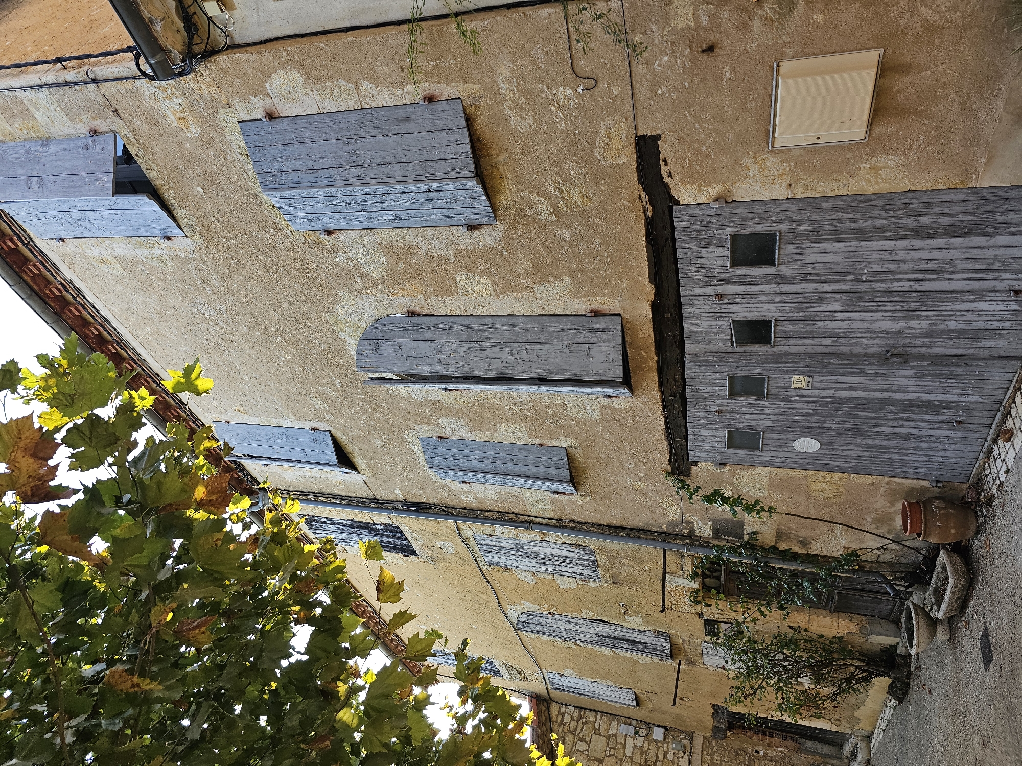 This is a low-angle, medium shot of a weathered, mustard-yellow building facade in Lavardens, France. The building has a rough, textured plaster finish over stone. Several weathered, gray wooden shutters are visible on the upper floors; some are partially open, revealing dark interiors. A large, gray wooden garage door dominates the lower portion of the building. This door features three small, square windows near the top. To the left of the garage door, a narrow alleyway is visible, with a dark wooden door and a small, bare-branched shrub in a concrete planter. In front of the garage door and planter, there are two more planters: a round, aged stone one and a rectangular concrete one, both containing soil or dried leaves. A large, dark brown clay pot sits to the right of the concrete planter, next to the garage door. Green vines are growing up the side of the building, particularly around a downspout that runs vertically along the facade. The leaves of a tree with green and yellow foliage hang from the upper left of the frame, partially obscuring the building. The overall impression is of an old, rustic structure in a European town.