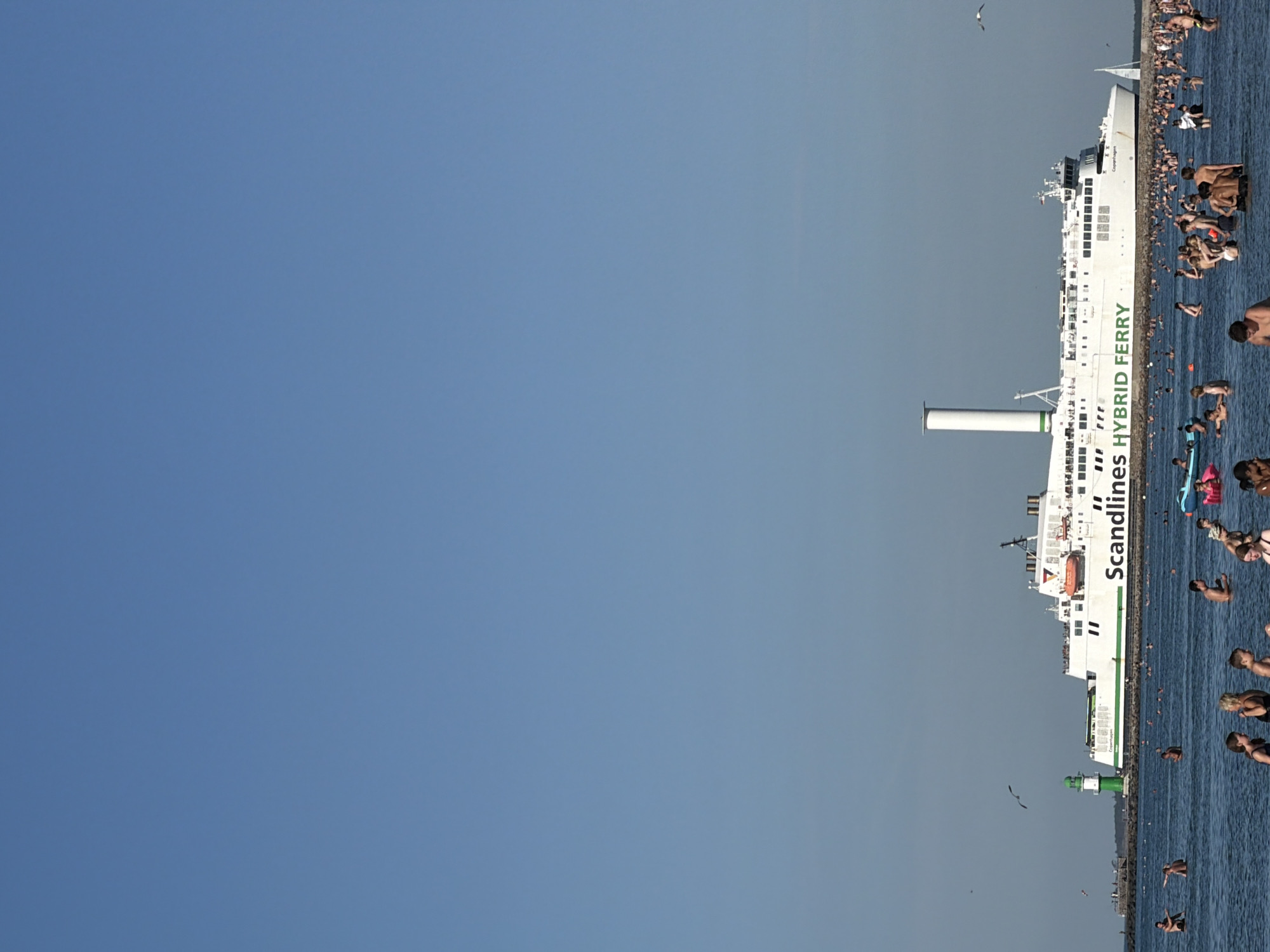 The image captures a sunny day at a beach in Rostock, Germany, with a large ferry prominently docked in the distance. The sea is populated with numerous swimmers and sunbathers enjoying the water. The ferry, identified as a "Scandlines HYBRID FERRY," is white with green accents and displays "Copenhagen" on its side. A green and white lighthouse stands near the ferry's docking area. The sky is a clear, vibrant blue.