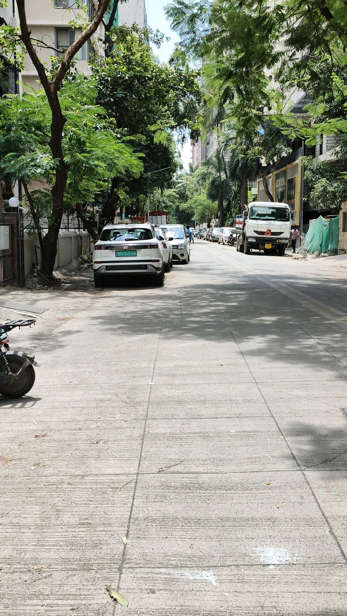 The image shows a street scene in Mumbai, India, likely in a residential or commercial area with buildings lining one side and parked vehicles on the other. The foreground is dominated by a concrete road with visible expansion joints and some debris. On the left side of the road, a white SUV is parked, bearing the license plate MH02GJ5343. Further down the street, several other cars and a white truck are parked or in motion. Tall green trees with dense foliage are prevalent along the sidewalk and between buildings, casting shadows on the road. The sky is partially visible at the top, suggesting it is daytime with a clear or partly cloudy sky. A person can be seen walking on the right side of the road near a green tarpaulin. The overall impression is that of a typical urban street environment in Mumbai during the day.