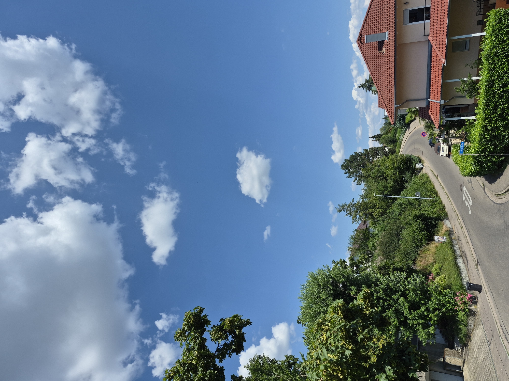 The image shows a residential street scene under a bright blue sky dotted with fluffy white clouds. The perspective is from a high vantage point, looking down a curved road lined with lush green trees and shrubs. On the right side of the road, a house with a red tiled roof and beige exterior is visible, with a neatly trimmed hedge bordering its property.
A white car is parked on the side of the road, and a street sign is visible. The road itself has markings, including a "30" indicating a speed limit. The overall impression is one of a peaceful, suburban setting on a clear, sunny day, likely in the late morning or early afternoon given the bright light. The greenery suggests a temperate climate. There are no people visible in the image. The scene evokes a sense of calm and everyday life.