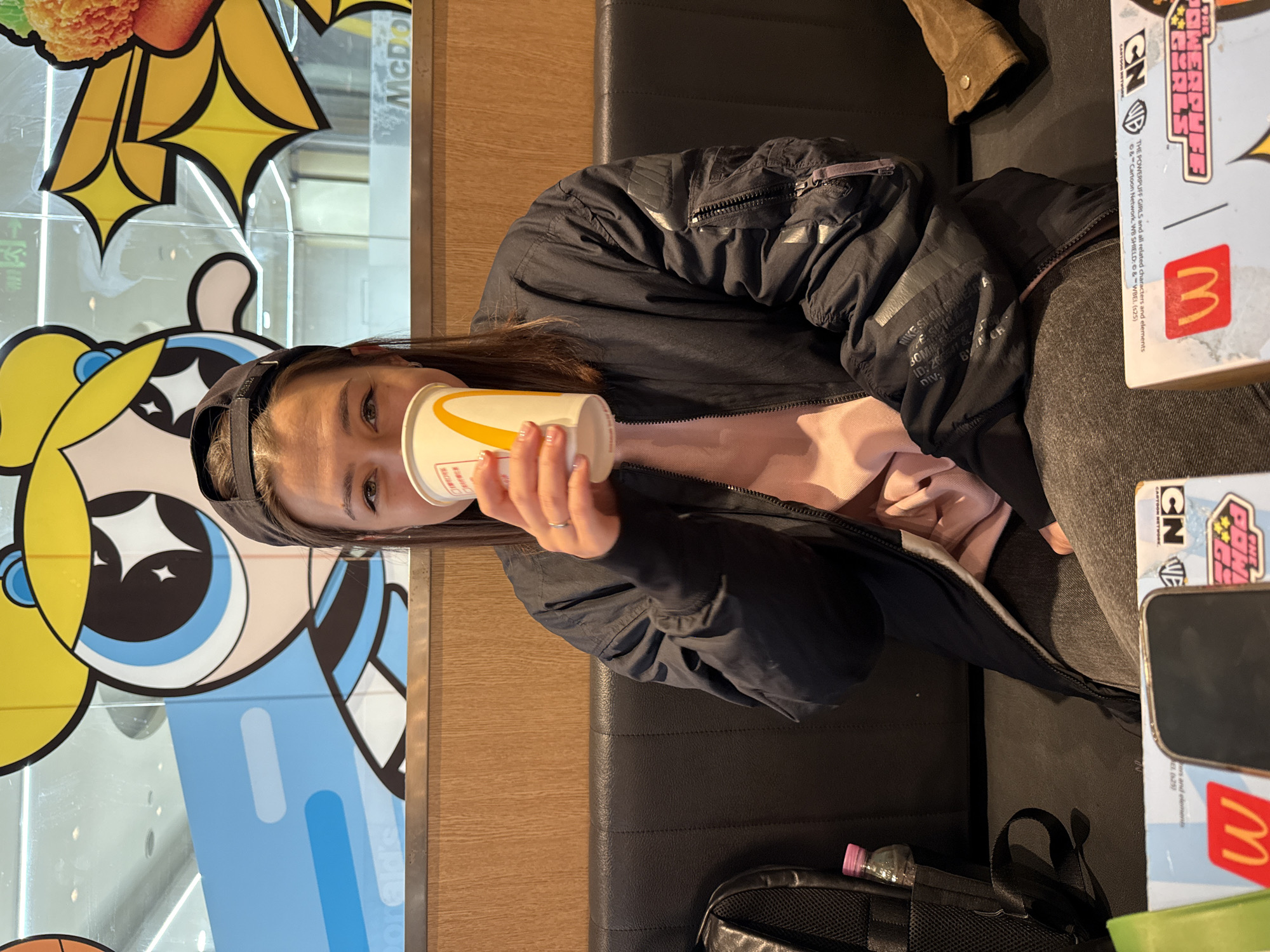 A woman with brown hair and wearing a black jacket and pink shirt is drinking from a yellow and white McDonald's cup. She is sitting on a black leather bench inside a McDonald's restaurant. Next to her on the bench is a phone, Powerpuff Girls McDonald's box and a backpack containing a water bottle on the floor. A Powerpuff Girls advertisement is visible on the window behind her. The photo was possibly taken in Dongcheng District, China.