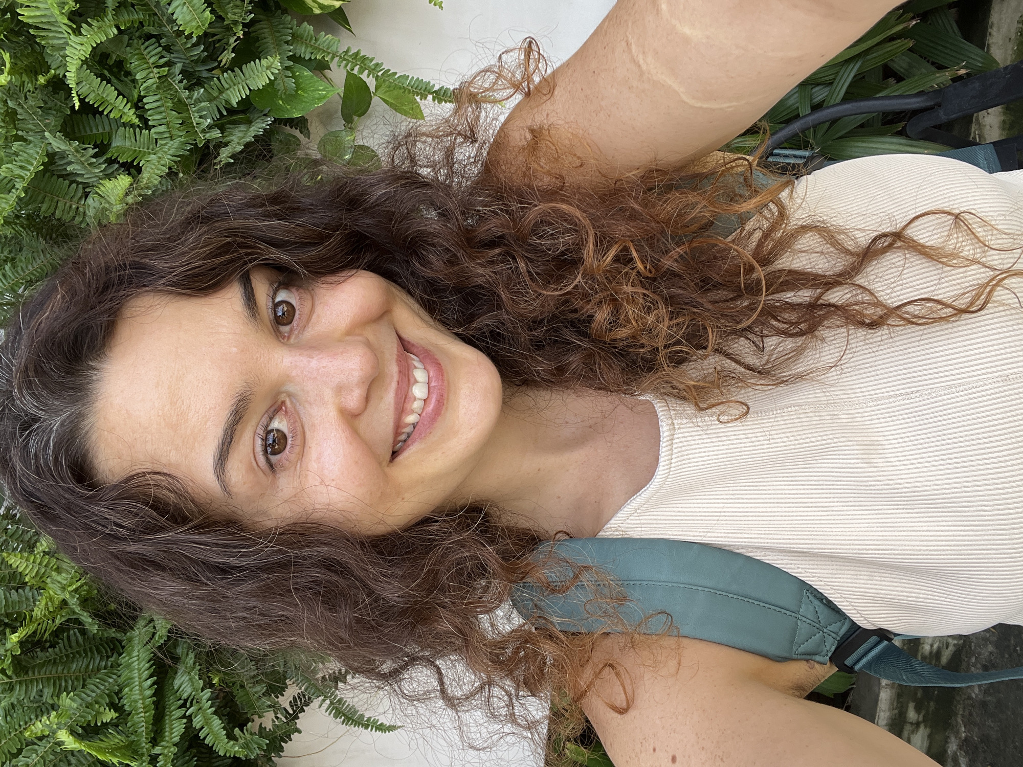 A woman with long, curly brown hair is taking a selfie in front of a wall covered in green ferns. She is wearing a light-colored, ribbed tank top and a teal backpack. Her eyes are brown, and she is smiling. The lighting suggests it is daytime. The background indicates an outdoor setting, likely in a tropical or subtropical environment. Given the presence of ferns and the overall ambiance, the location could be a garden or a courtyard in Mũi Né, Vietnam.