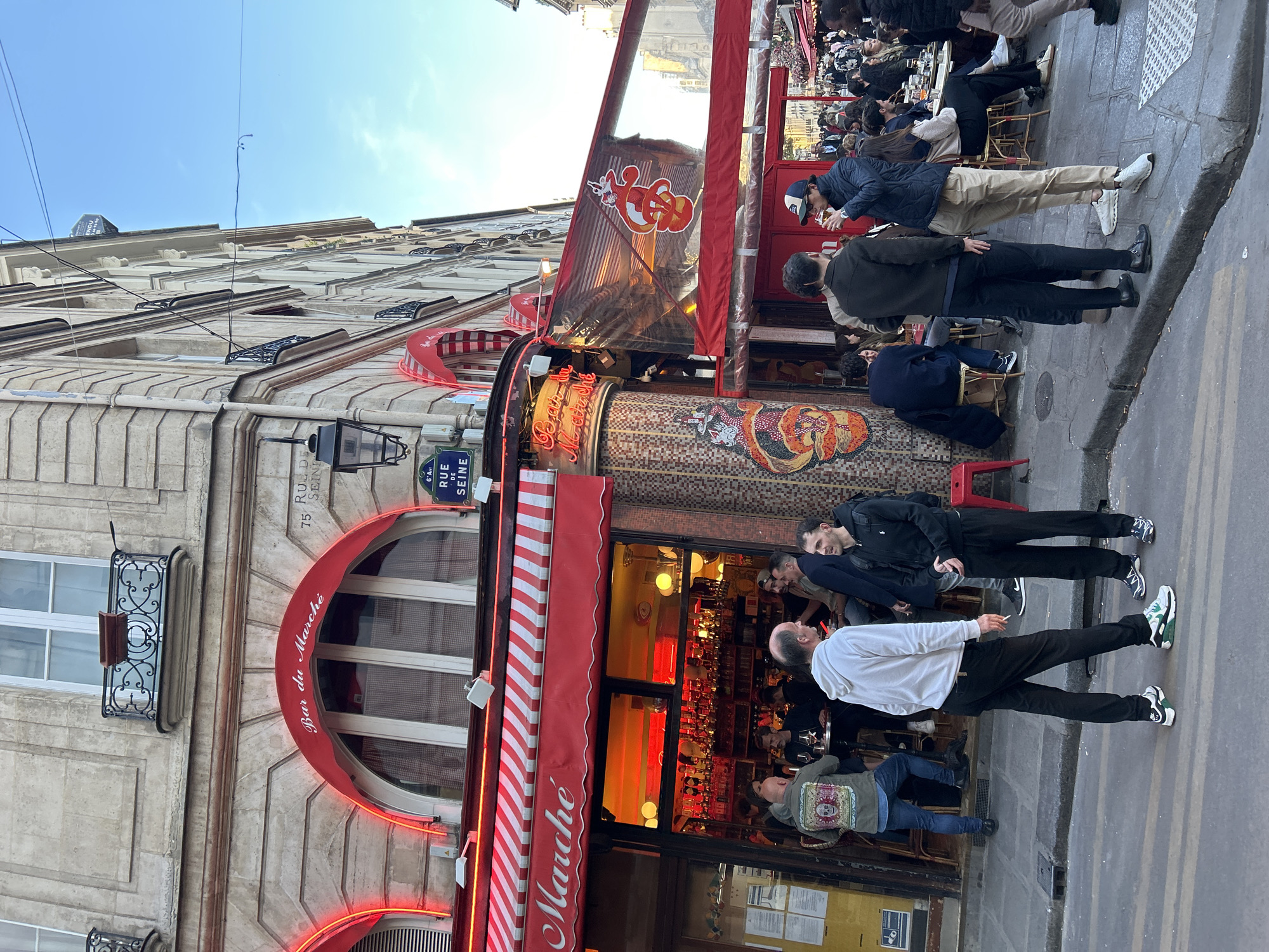The image depicts a lively street scene in front of a bar or café, likely in Paris, given the visible street name "Rue de Seine". The setting is daytime, under a clear blue sky, with the sun casting some shadows.
The main subject is a multi-story building featuring classic European architecture with light-colored stone facades, ornate balconies, and large windows. The ground floor houses a bar, identifiable by its prominent red awnings, neon lighting, and visible interior. A large red and white striped awning extends over the entrance, with "Bar du Marché" written in white cursive text. Another arched red awning also displays "Bar du Marché". To the right of the entrance, an extended red canopy or windbreak features a distinctive red logo of two intertwined, stylized creatures, possibly serpents or dragons. A decorative tiled pillar beneath this canopy also features similar serpentine mosaic patterns.
Numerous people are gathered outside the establishment. Several individuals are standing on the paved sidewalk, some appearing to be socializing or waiting. Two men are prominent in the foreground, one in a white long-sleeve shirt holding what appears to be a cigarette, and another in a black jacket and black pants. Others are seated at tables under the outdoor canopy, while more patrons are visible inside the bar through its windows, with bottles illuminated behind the counter. The overall activity suggests a social gathering at a popular establishment.
Notable details include the building number "75" visible above an arched window. A blue and green street sign reads "6e Arr. Rue de Seine". In the distant background, high on another building, a faint "VENDU" (Sold) sign can be seen. The general atmosphere is bustling and casual.