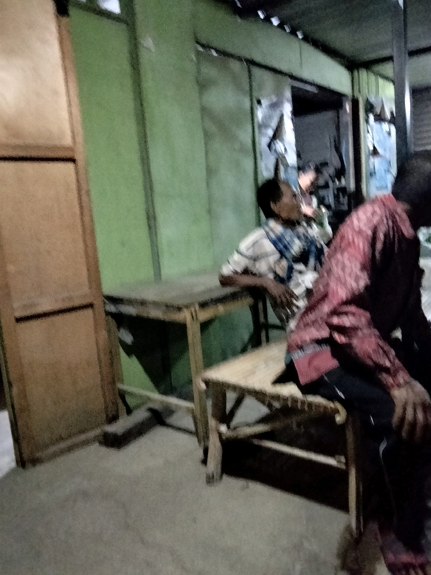 In Mrican, Indonesia, two men are seated at a wooden table. The man on the left wears a checkered shirt and is looking to his right. The man on the right is partially obscured and wears a red patterned shirt. They are positioned in an outdoor or semi-outdoor environment with green walls and a concrete floor. A wooden door is visible on the left side of the frame.
