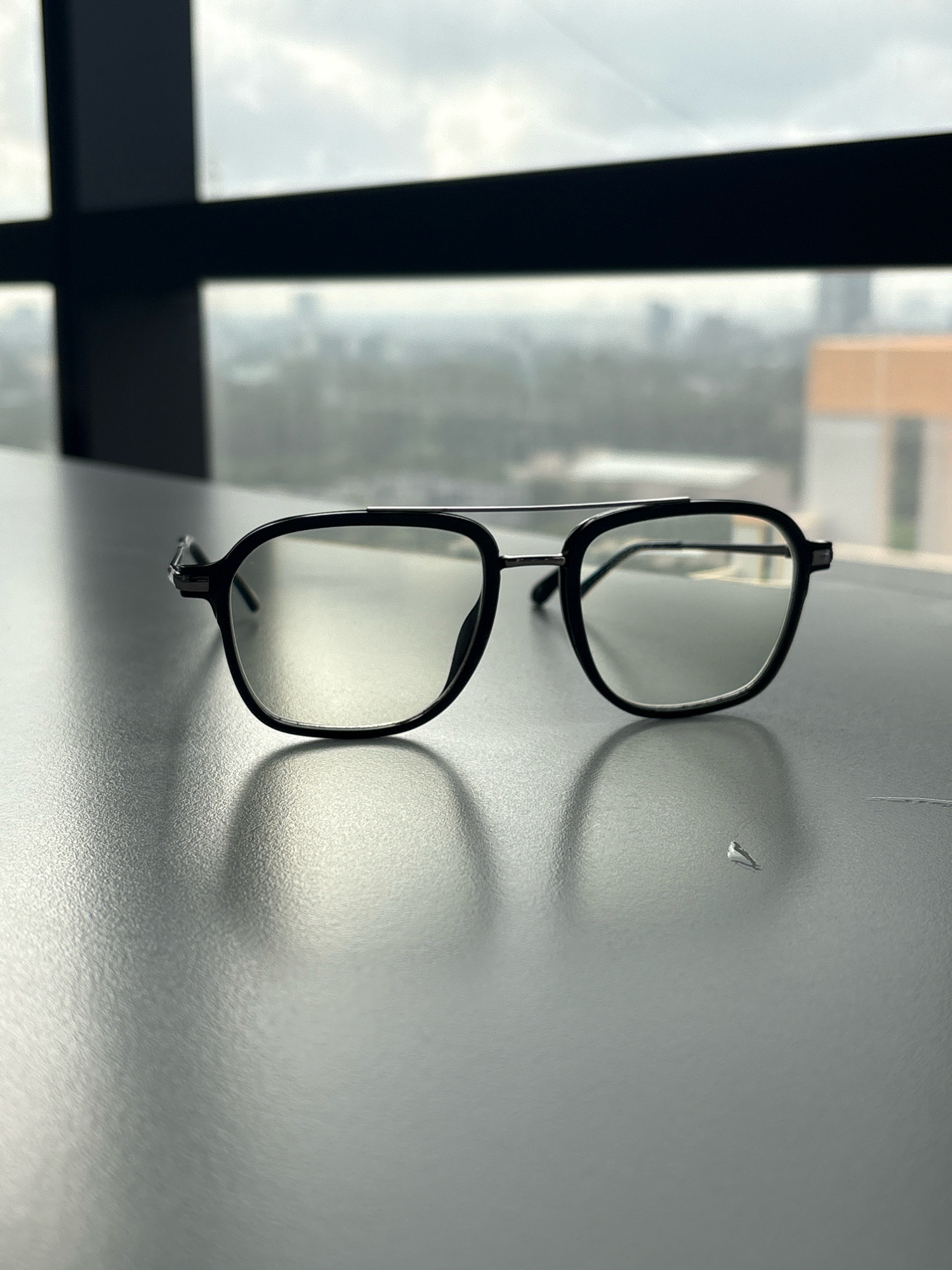 A pair of black-rimmed eyeglasses with clear lenses and a silver bridge rests on a dark gray, textured surface. The surface appears to be a table or desk. In the background, out of focus, is a blurred cityscape seen through a window with dark vertical and horizontal dividers. The sky appears overcast. The eyeglasses are positioned centrally in the frame, slightly angled towards the left. A subtle shadow is cast by the eyeglasses onto the surface. The scene is indoors, possibly in an office or apartment with a view of the city of Pune, India, indicated by the presence of buildings and infrastructure in the distant, blurred background.