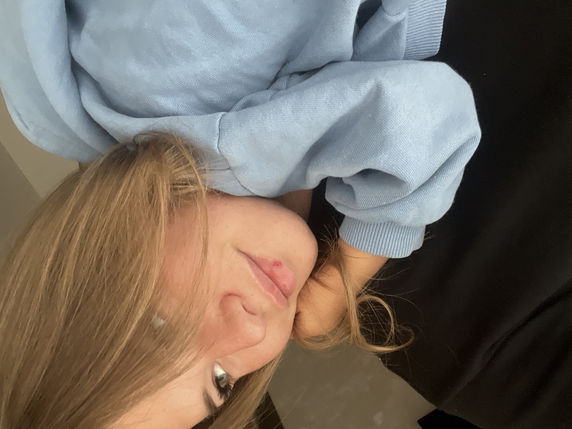 A close-up, rotated image shows a young woman with light brown hair and light skin. She is wearing a light blue sweater. She is partially reclined, with her head resting on her hand or arm. A small blemish or cut is visible on her upper lip. A dark, possibly black, fabric is present in the background. There is no indication of the location being in Budva, Montenegro, or any other specific location.