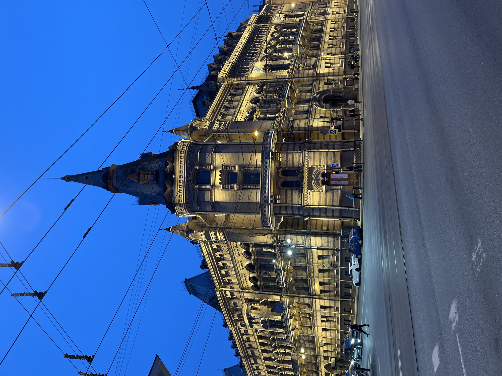 The image depicts a street scene at dusk or night in Saint Petersburg, Russia, centered on the illuminated Singer House (Dom Knigi) located at Nevsky Prospect, 28. The multi-story building, an example of Art Nouveau and eclectic architecture, features a light-colored facade with darker horizontal stripes, adorned with numerous arched windows, small balconies, and ornate detailing. A prominent corner tower with a distinct spire rises towards the deep blue twilight sky.
Overhead, a complex network of black electrical wires, likely for public transport like trams or trolleybuses, spans the upper portion of the frame. The wide asphalt road in the foreground shows faint white markings.
On the left side of the road, a dark-colored SUV is partially visible with its headlights on. Further right, a white sedan and a blue sedan are parked or stopped near the curb in front of the building.
Approximately 10-12 individuals are present. One person in dark clothing walks left-to-right across the road in the mid-left. Several other pedestrians are visible walking or standing on the illuminated sidewalks along the building's base, particularly on the right side.
Streetlights and traffic signals, emitting warm yellow light, are visible on the sidewalks and at the intersection. A black metal fence or railing runs parallel to the sidewalk on the left, and a yellow diamond-shaped road sign is affixed to the building's facade.