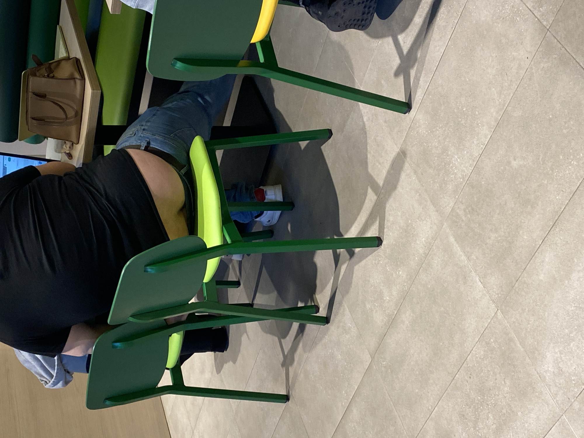 A person wearing a black shirt and jeans is seated at a table with a tan bag on it. They are sitting in a green chair with a yellow back. Next to them is another green chair with a green back, seemingly stacked. Their legs are visible, and they are wearing white sneakers with red accents. The floor is tiled with light-colored, square tiles. The scene appears to be indoors. The location in Varna, Bulgaria, is undetermined from the provided visual data.