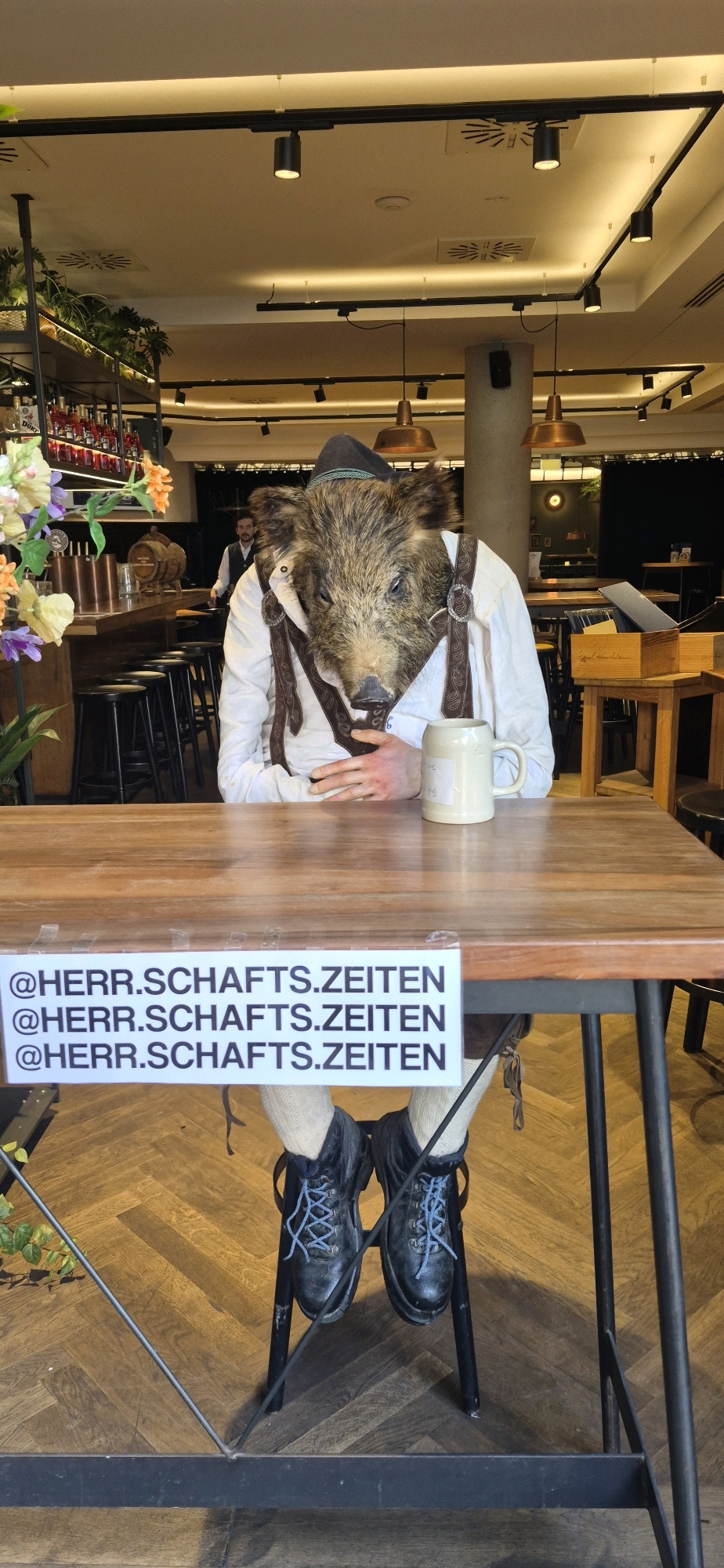 The image shows a display of a person wearing a boar mask and traditional Bavarian clothing, sitting at a wooden table in what appears to be a restaurant or beer hall. The person is wearing a white shirt, brown suspenders, and a hat. They are holding a beer mug. In front of the table, a sign with the text "@HERR.SCHAFTS.ZEITEN" repeated three times is attached. The background shows typical restaurant seating, lighting, and decor, including a bar area with bottles and a keg. The flooring is a herringbone pattern. Based on the signage and attire, this scene is likely located in Munich, Germany, a city known for its beer culture and traditional Bavarian customs.