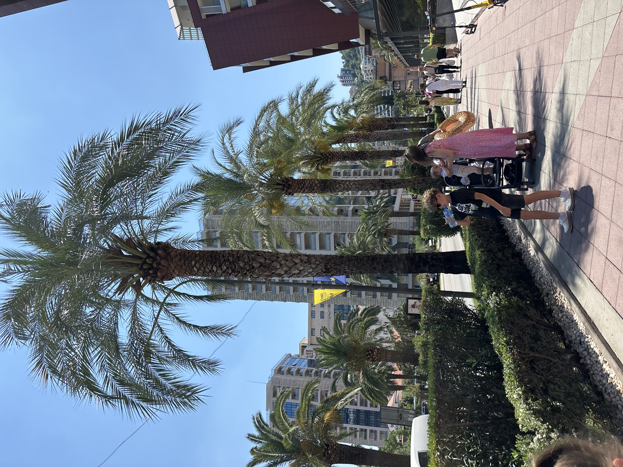 The image depicts a street scene in Budva, Montenegro, on a sunny day. In the foreground, a young boy wearing a black t-shirt and shorts drinks from a water bottle while standing next to a stroller. A woman in a pink dress is beside the stroller, holding a baby. Behind them, several people are walking along a tiled sidewalk lined with palm trees. The background features modern buildings and a hillside with more architecture. The environment is a well-maintained urban area with lush greenery and clear blue skies.