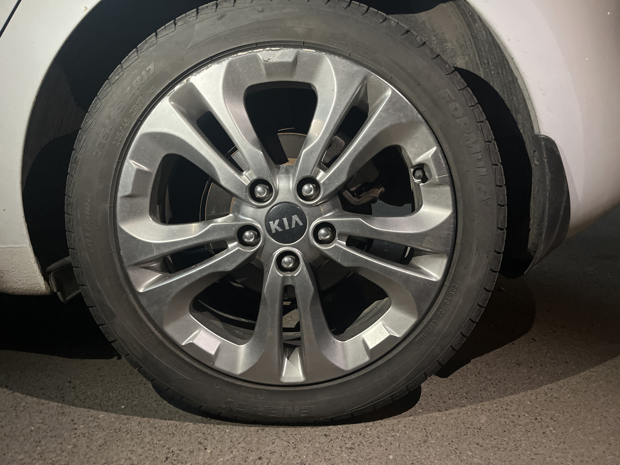 The image shows a close-up of a car tire. The tire is black and has the word "ENERGY" visible on the sidewall, along with the size specification "225/45 ZR17 94Y." The wheel is silver with a multi-spoke design and a "KIA" logo in the center. The car appears to be white, with part of the body visible above and to the side of the tire. The tire is resting on asphalt, indicating it is likely parked on a road or parking lot. The lighting suggests the image was taken in the evening or under artificial light. There is no information available in the image to specify the location as Kazan, Russia.