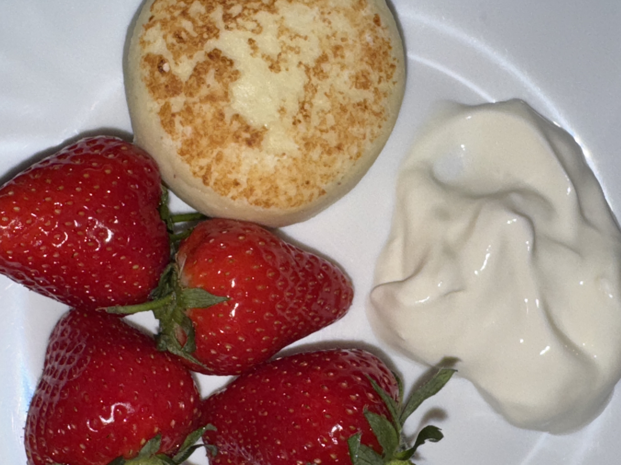 A white plate contains four strawberries with green stems, a round, browned pancake or syrniki, and a dollop of white cream. The location and city of Moscow, Russia cannot be ascertained.