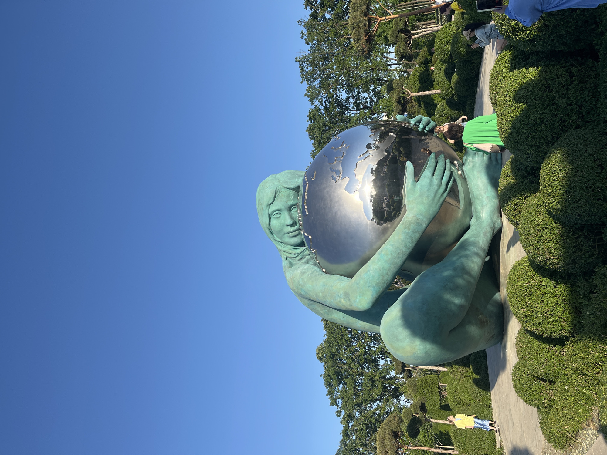 A photograph captures a statue in a park in Krasnodar, Russia. The statue, resembling a seated female figure, is made of a green material and holds a reflective globe. The environment surrounding the statue is green and lush, including meticulously manicured bushes. The sky is blue, and there are several people in the background, some standing and walking. A woman in a yellow jacket is visible near the base of the statue. The photograph is taken at an angle, tilting the composition.