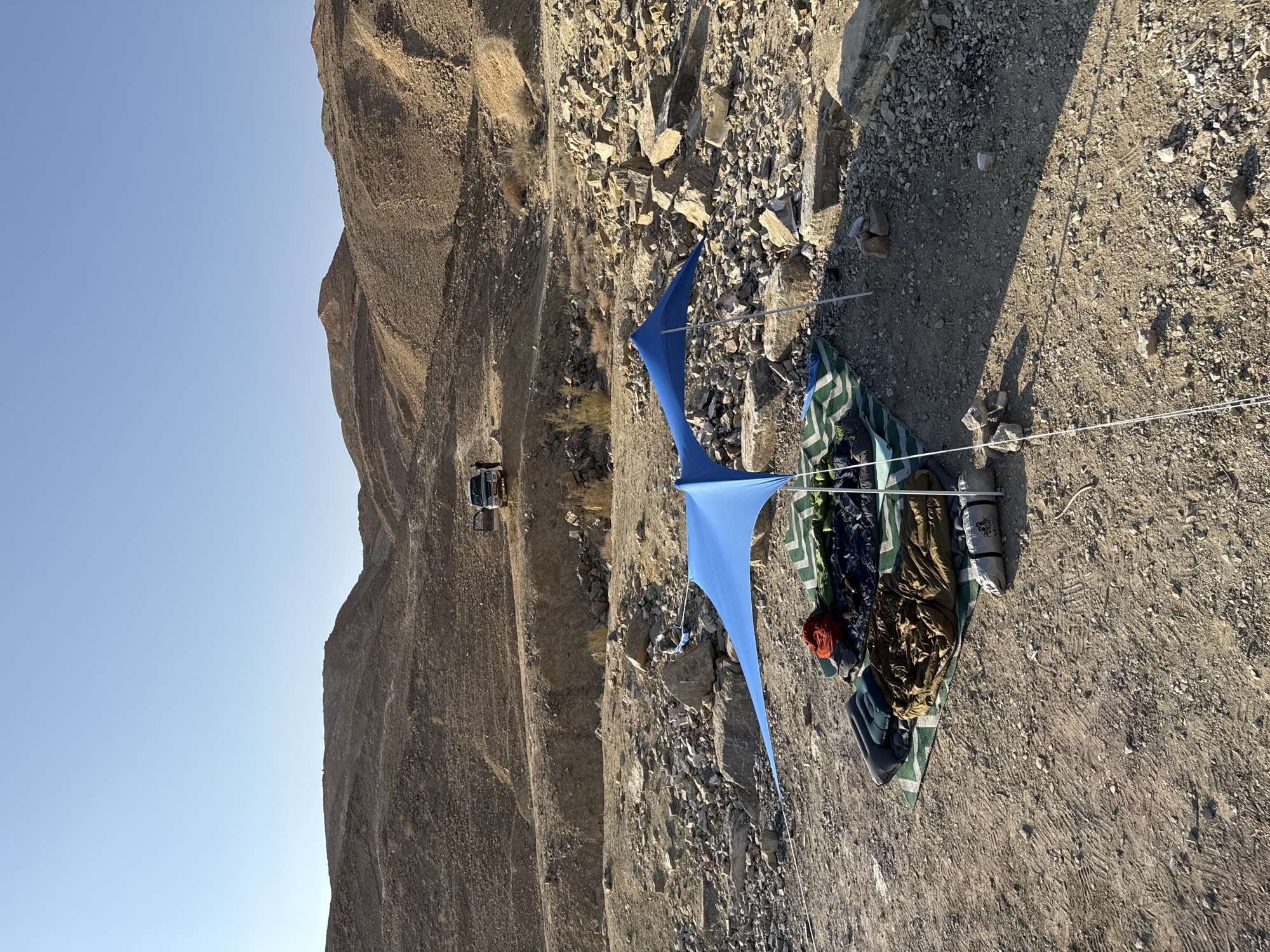 The image depicts a campsite set up in a desert landscape within the Ramat Negev Regional Council, Israel. The ground is composed of dry soil, gravel, and rocks. In the foreground, a blue canopy tent is erected, providing shade over a green and white striped mat. Several sleeping bags, an inflatable mattress, and a red bag are spread out on the mat. A rolled-up tent is also present. In the background, a dirt road winds through barren, rocky hills under a clear blue sky. A silver SUV with its doors open is parked on the road, further up the incline. The overall scene suggests a remote outdoor recreational activity.