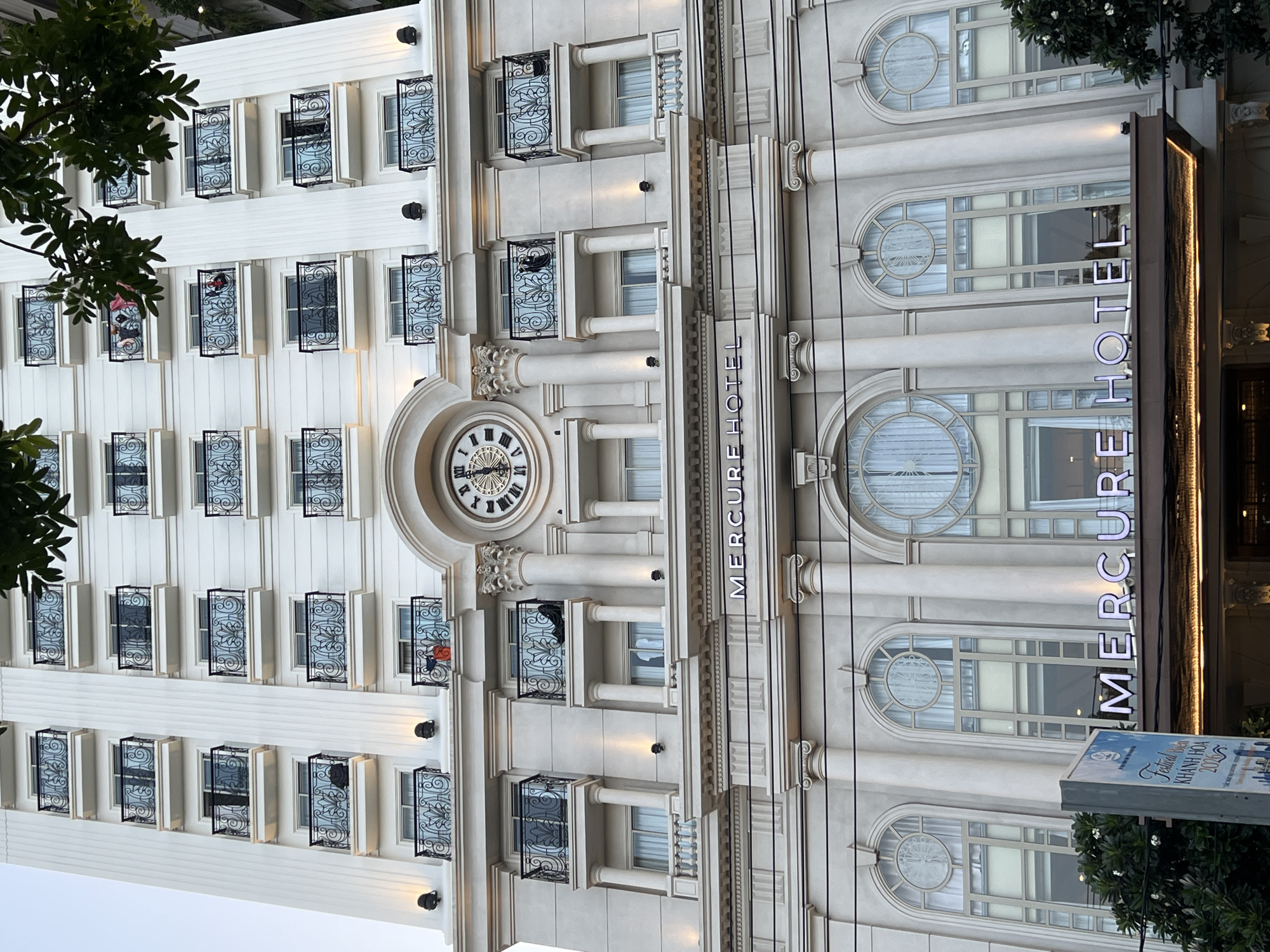 The image shows the Mercure Hotel Nha Trang, a tall, white building with multiple floors and balconies. The hotel features classical architectural elements, including columns and ornate railings on the balconies. A large clock face is visible on the facade of the building. The hotel's name, "MERCURE HOTEL," is illuminated with neon lights above the entrance. In the foreground, a signpost with Vietnamese text and graphics is partially visible, and trees frame the bottom of the image. The sky is overcast, suggesting it is either early morning or late afternoon.