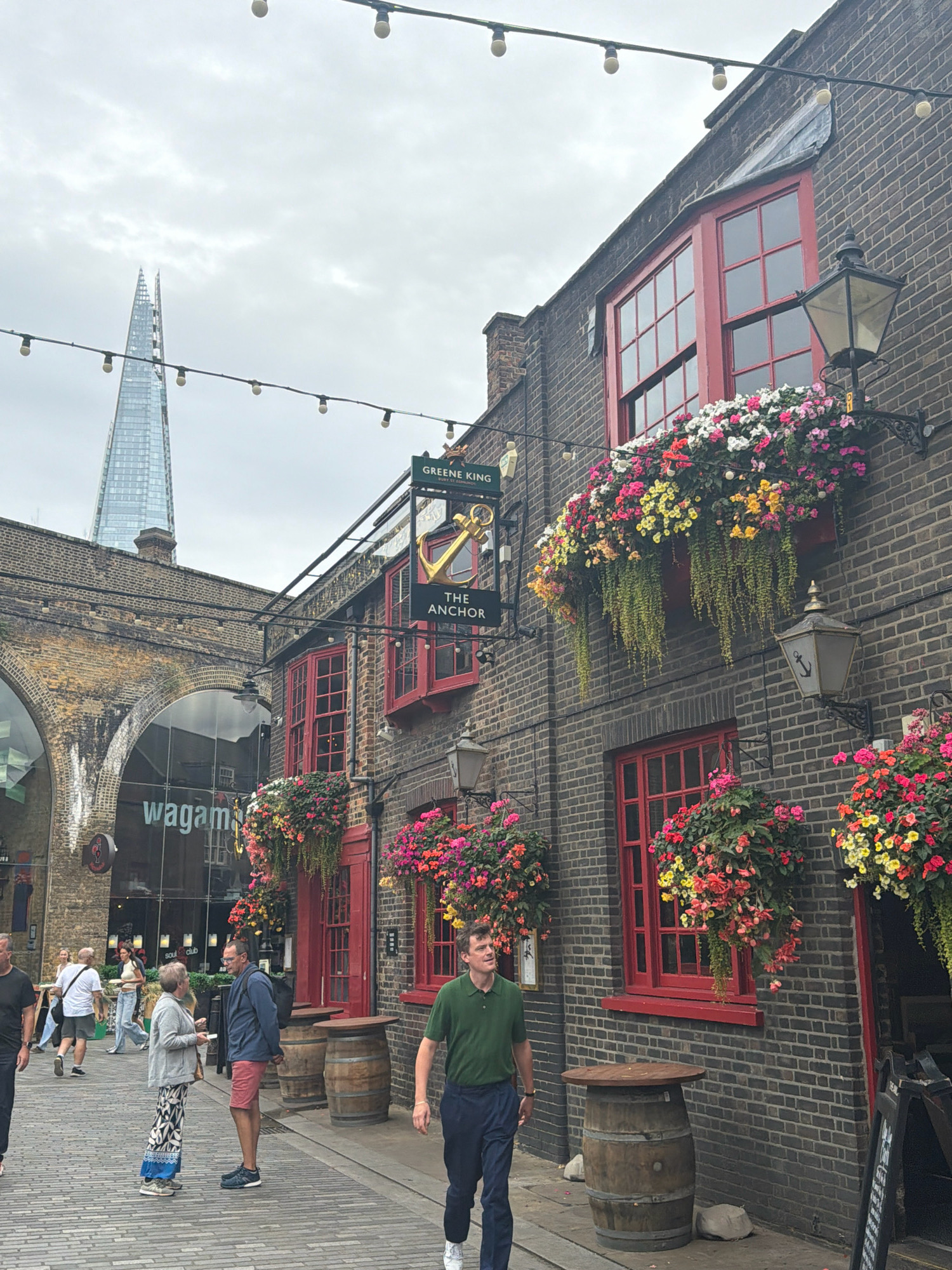 This is a medium shot, eye-level, outdoor photograph taken on a cloudy day. The scene depicts a street in London, United Kingdom, likely a tourist area given the prominent presence of "The Shard" skyscraper in the background.
In the foreground and midground, a narrow cobbled street runs towards the right of the frame. On the right side of the street is a brick building with red-trimmed windows. This building has a sign that reads "THE ANCHOR" with "GREENE KING" above it. Hanging baskets filled with colorful flowers, including pink, yellow, and white blooms, adorn the facade of the building above the windows and doorways. Wooden barrel tables are placed outside the establishment. Two wooden barrels stand to the left of the building entrance.
Several people are present on the street. On the left side of the street, two men are walking away from the camera. To the right of them, a woman in a light-colored top and jeans is walking towards the camera. Closer to the center, a man and a woman are engaged in conversation, facing each other. The man is wearing a dark blue jacket and red shorts, while the woman is wearing a grey cardigan and a patterned skirt. Further to the right, a man in a green polo shirt and dark trousers is walking towards the right side of the frame.
In the background, a large arched brick structure can be seen, with the "wagamama" restaurant logo visible through a glass facade. Beyond this structure, the distinctive glass skyscraper, "The Shard," pierces the sky. String lights with bulbs are suspended across the upper portion of the frame, contributing to the urban ambiance. The overall atmosphere is that of a bustling street scene in a historic part of London.