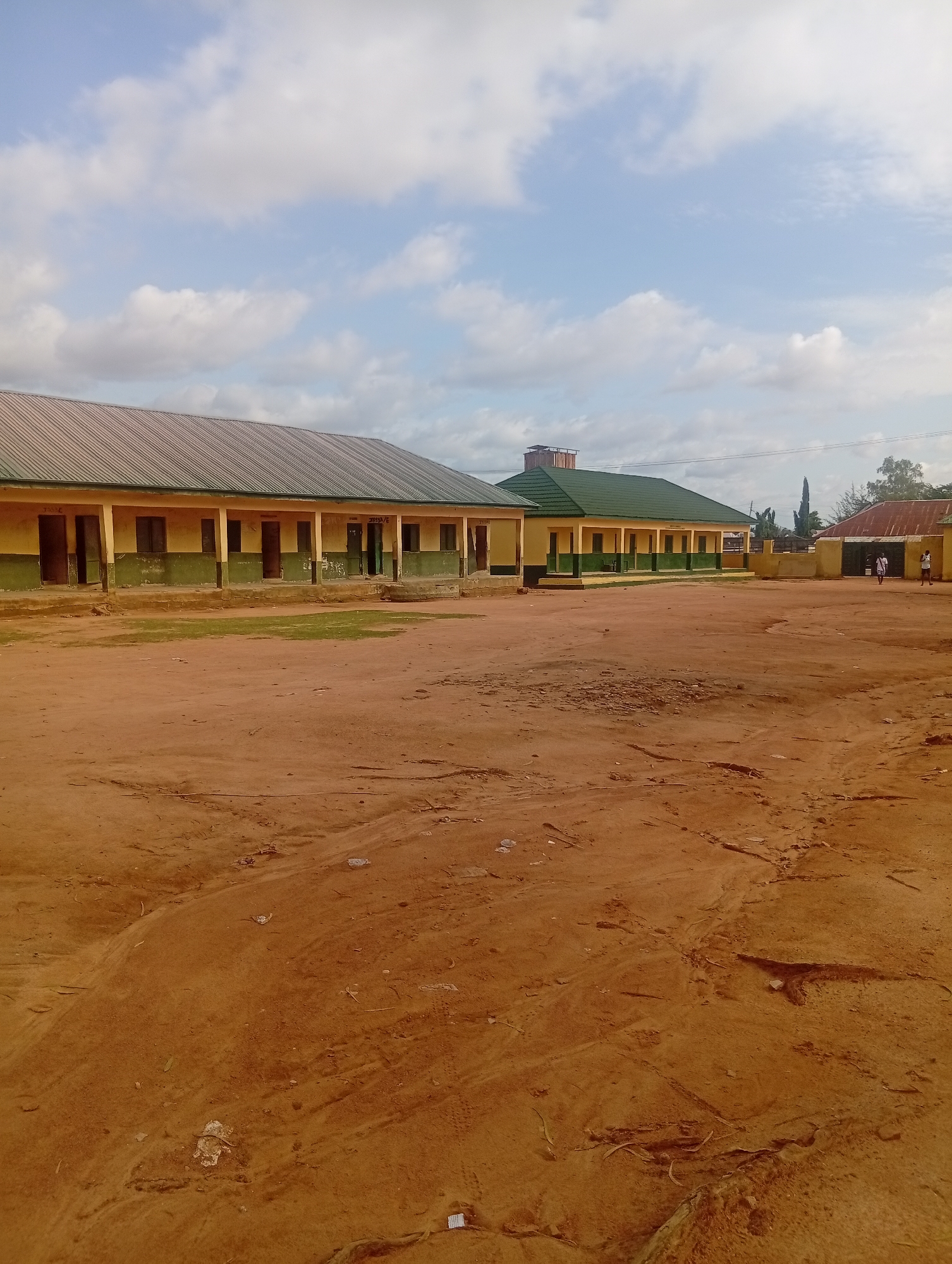 An outdoor scene in the city of Unknown, Unknown, depicts a compound with multiple single-story buildings and an extensive unpaved dirt ground. The sky above is light blue with scattered white cumulus clouds.
In the mid-ground, a long, single-story building on the left features a dark grey corrugated metal roof, light yellow walls, and a dark green lower section. Multiple open doorways are visible along its facade, with faint markings above some, such as "JSS3E," suggesting classroom designations. To its right, another single-story building has a dark green corrugated metal roof and similarly colored walls, with a prominent, possibly structural, element on its roof. Further to the right, a third building with a reddish-brown corrugated metal roof is partially visible, adjacent to a gate structure.
The foreground and much of the middle ground consist of reddish-brown, dry, unpaved earth, showing signs of vehicle tracks and footprints. Scattered debris, including small plastic items, paper scraps, and natural detritus, is present across the ground. Small patches of green vegetation are visible near the bases of the buildings.
Two individuals are discernible in the far right mid-ground, near the gate. One person, wearing a white top and dark bottoms, is walking towards the gate. Another person, also in light-colored clothing, stands near the gate.
Beyond the buildings on the right, several trees and shrubs are visible, including a distinct tall, slender tree. The scene is illuminated by daylight, indicating a time between morning and late afternoon.