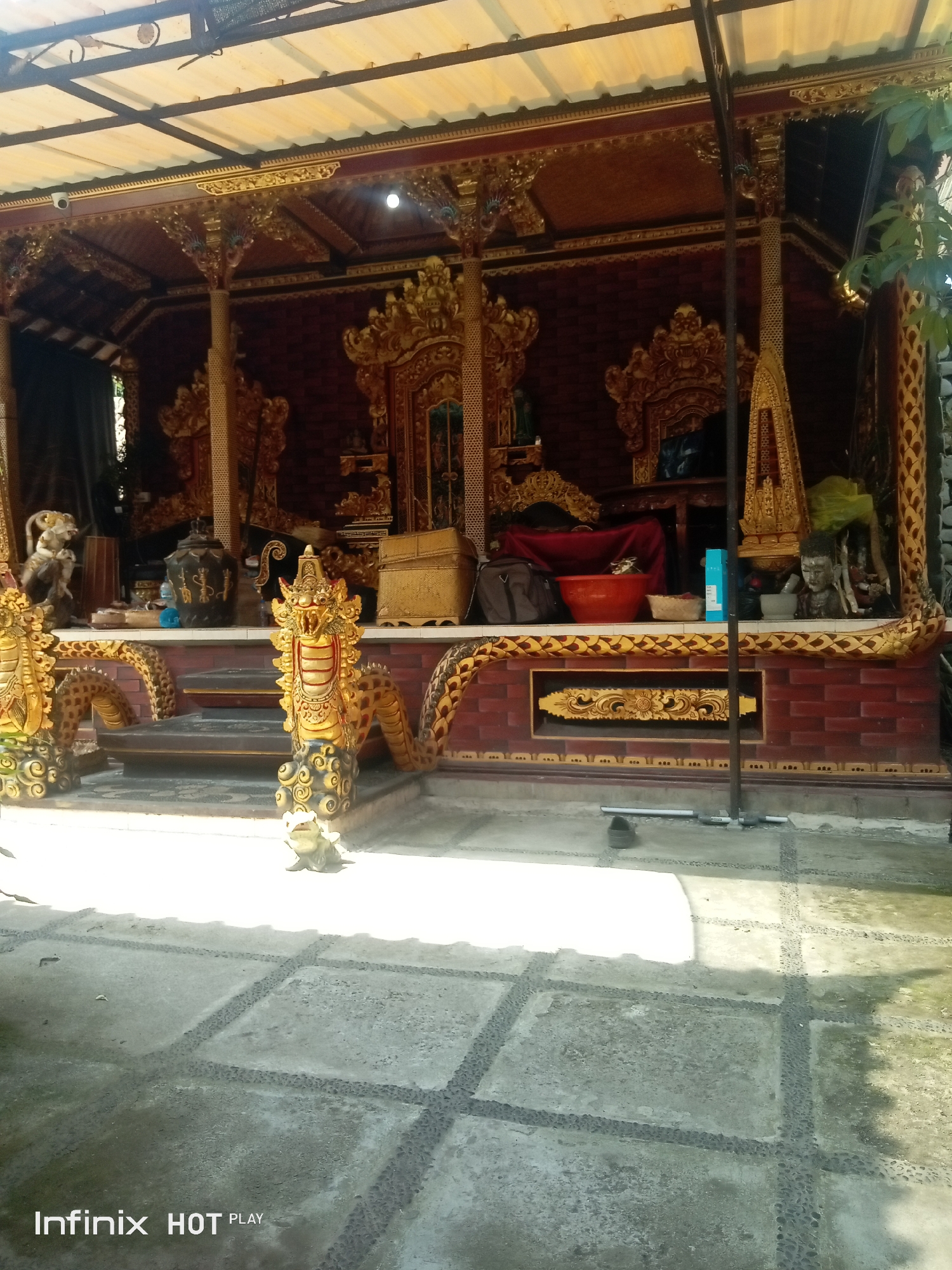 The image depicts a detailed view of a Balinese temple structure in Blahbatuh, Indonesia. The temple features ornate golden dragon carvings on pillars, intricate wood carvings along the facade and roof, and brickwork. A red cloth covers a platform at the rear, with several objects placed on shelves and surfaces, including ceramic pots, a woven basket, a red bowl, and sculpted heads. The foreground shows a tiled courtyard with a unique pattern of concrete squares bordered by small stones. The scene is illuminated by natural light, casting shadows on the ground. The overall impression is of a cultural and religious site with rich artistic detailing.